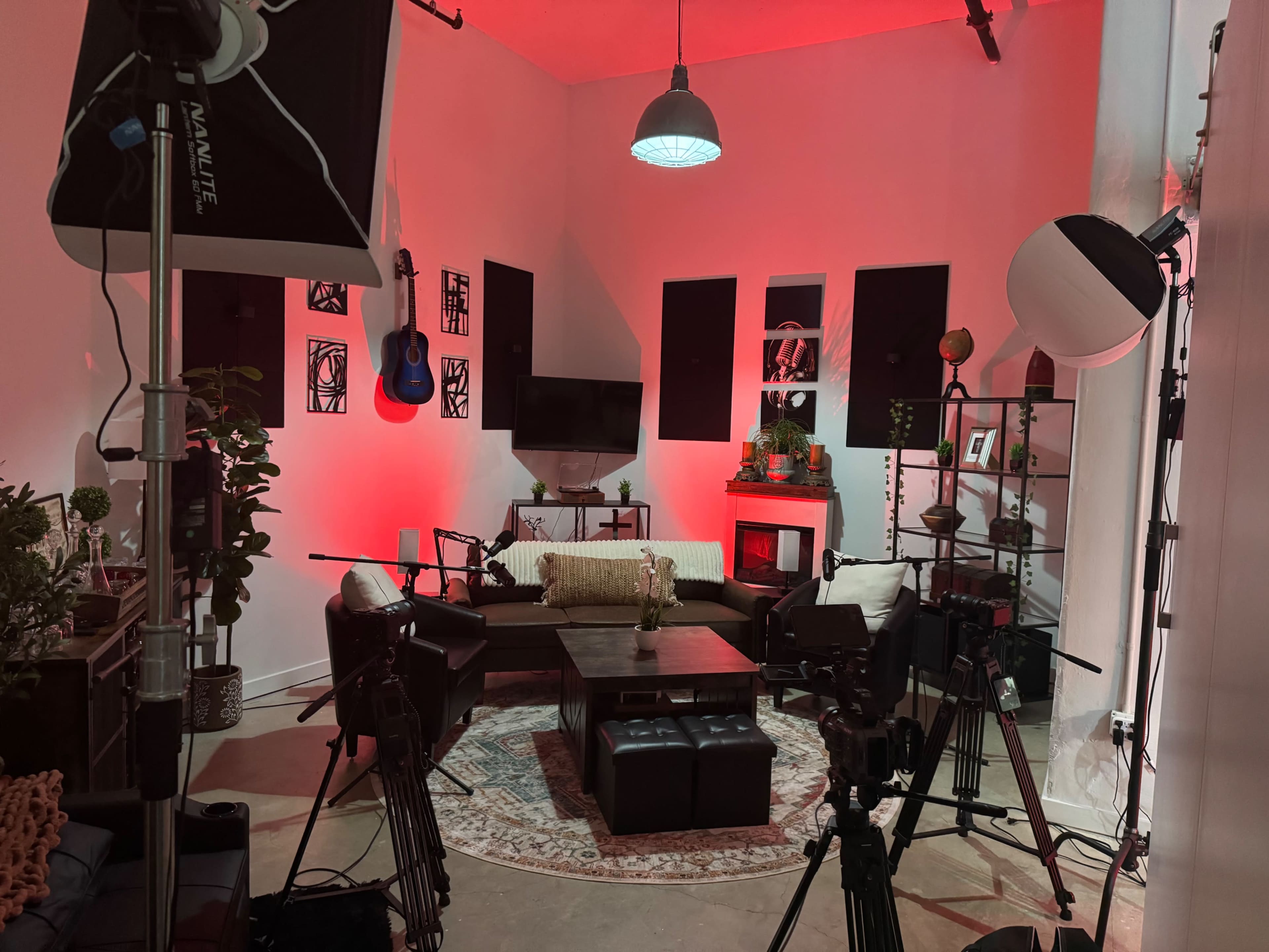 The image shows a studio setup with red lighting, featuring a cozy seating area, a coffee table, and multiple cameras positioned around the space.