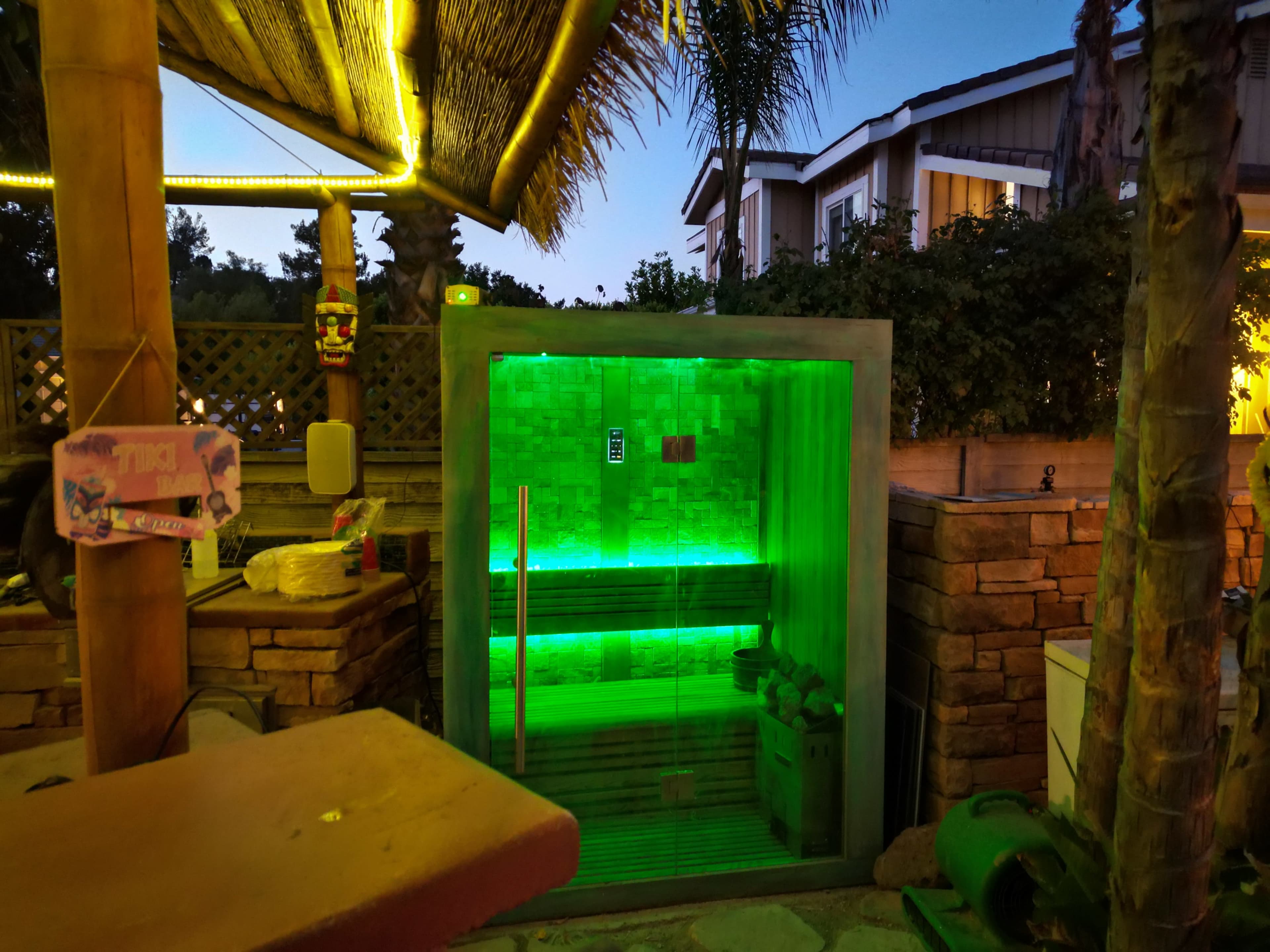 The image shows a modern sauna with green lighting, situated outdoors next to a stone wall and palm trees at dusk.