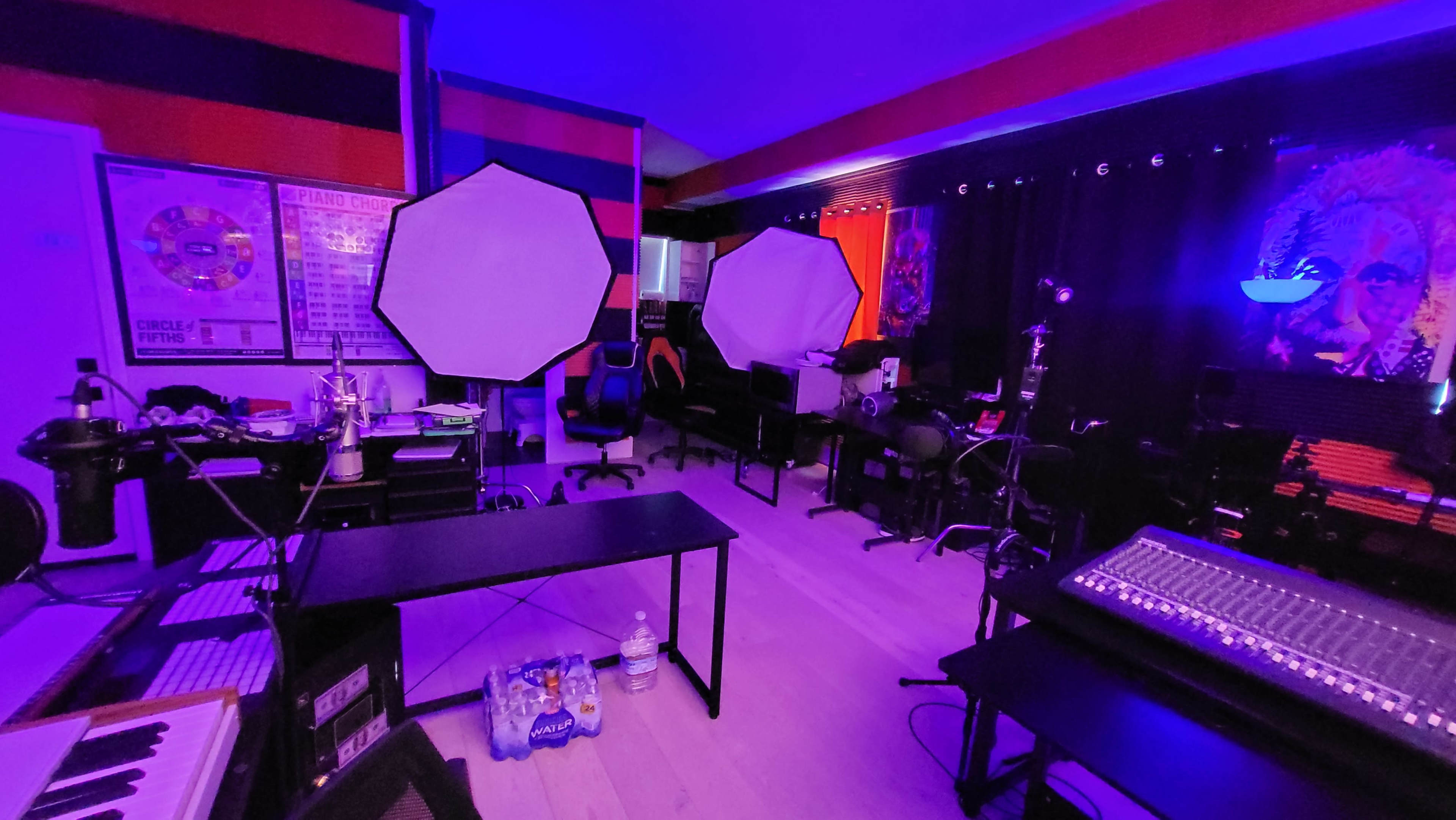 The image shows a dimly lit recording studio with two large softboxes, various musical equipment, and chairs arranged around a central table.