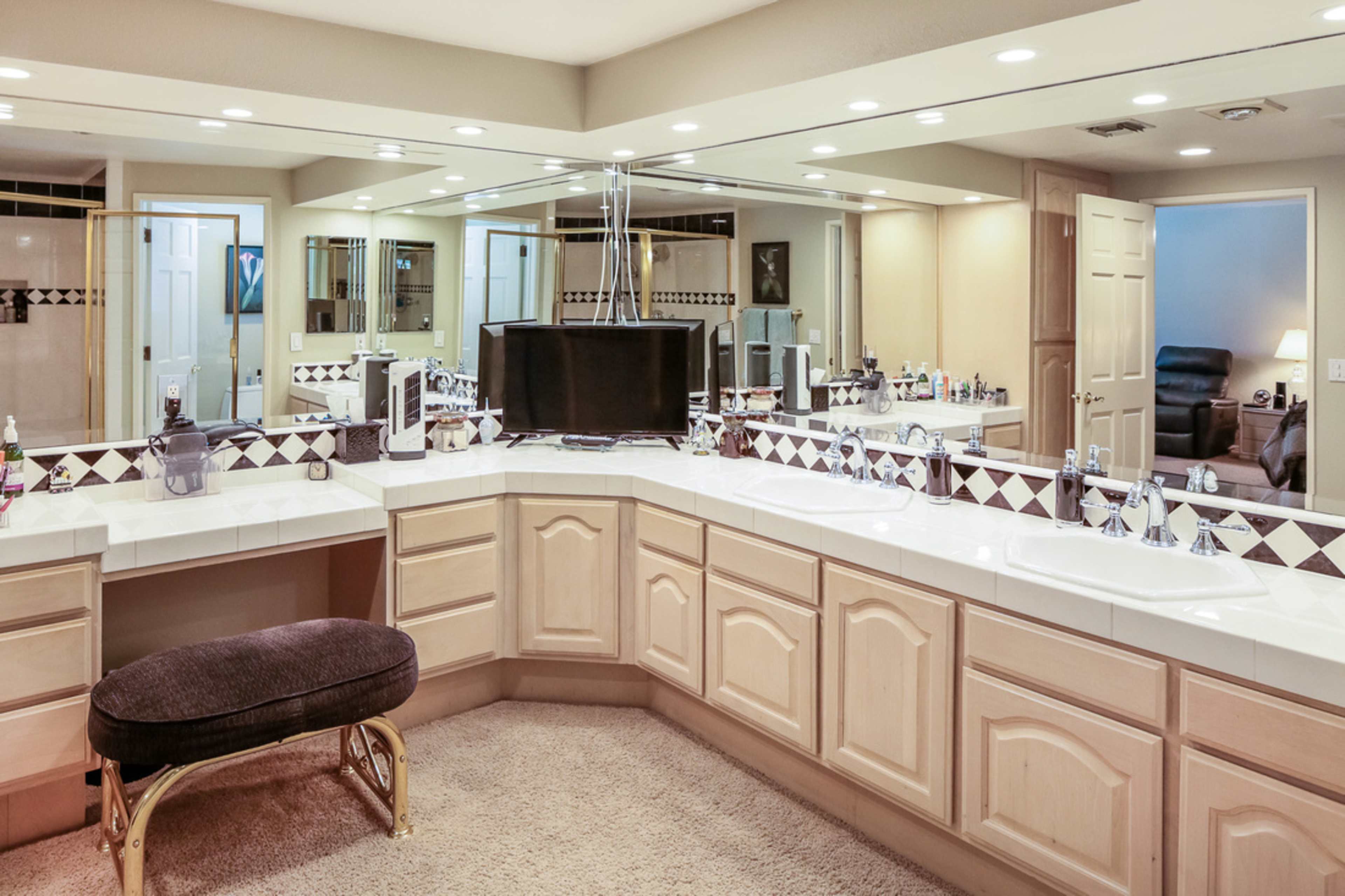 A spacious bathroom features dual sinks with mirrored cabinetry and a vanity area, complemented by a carpeted floor and various toiletries arranged on the countertops.
