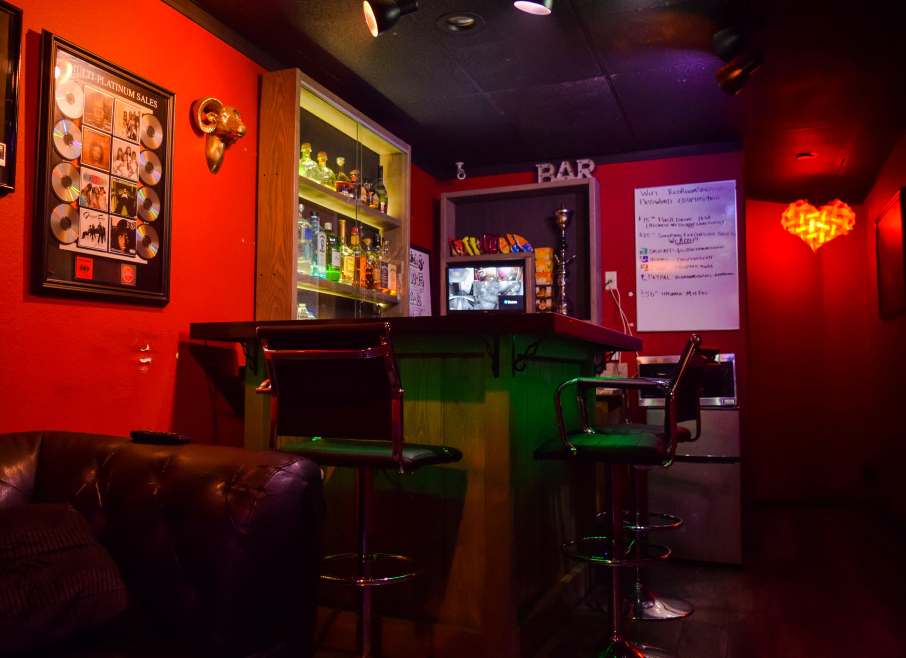 The image shows a cozy bar area with a wooden counter, shelves lined with bottles, and red walls adorned with framed art.