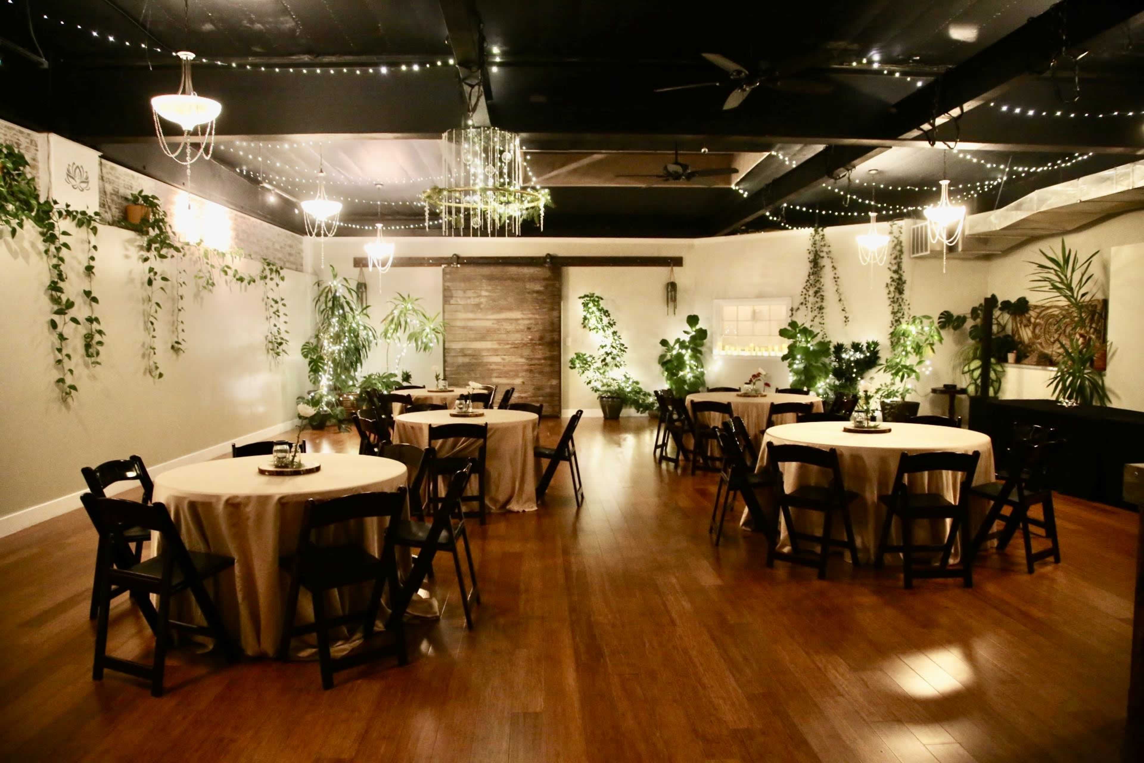 The room features several round tables covered with cream tablecloths, surrounded by black chairs, and is decorated with plants and dim lighting.