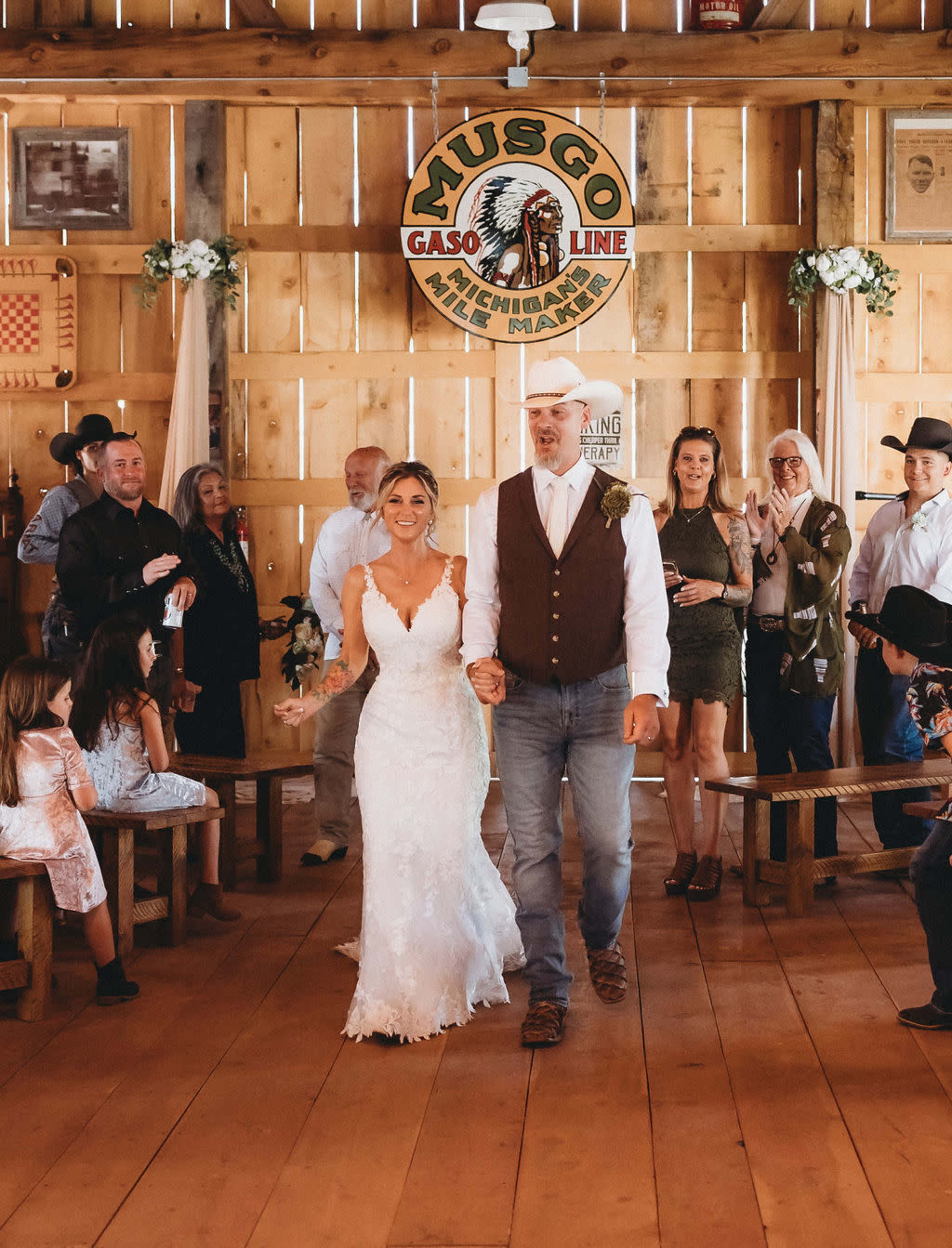 A bride and her father walk down a wooden aisle in a rustic venue, surrounded by guests seated on benches.