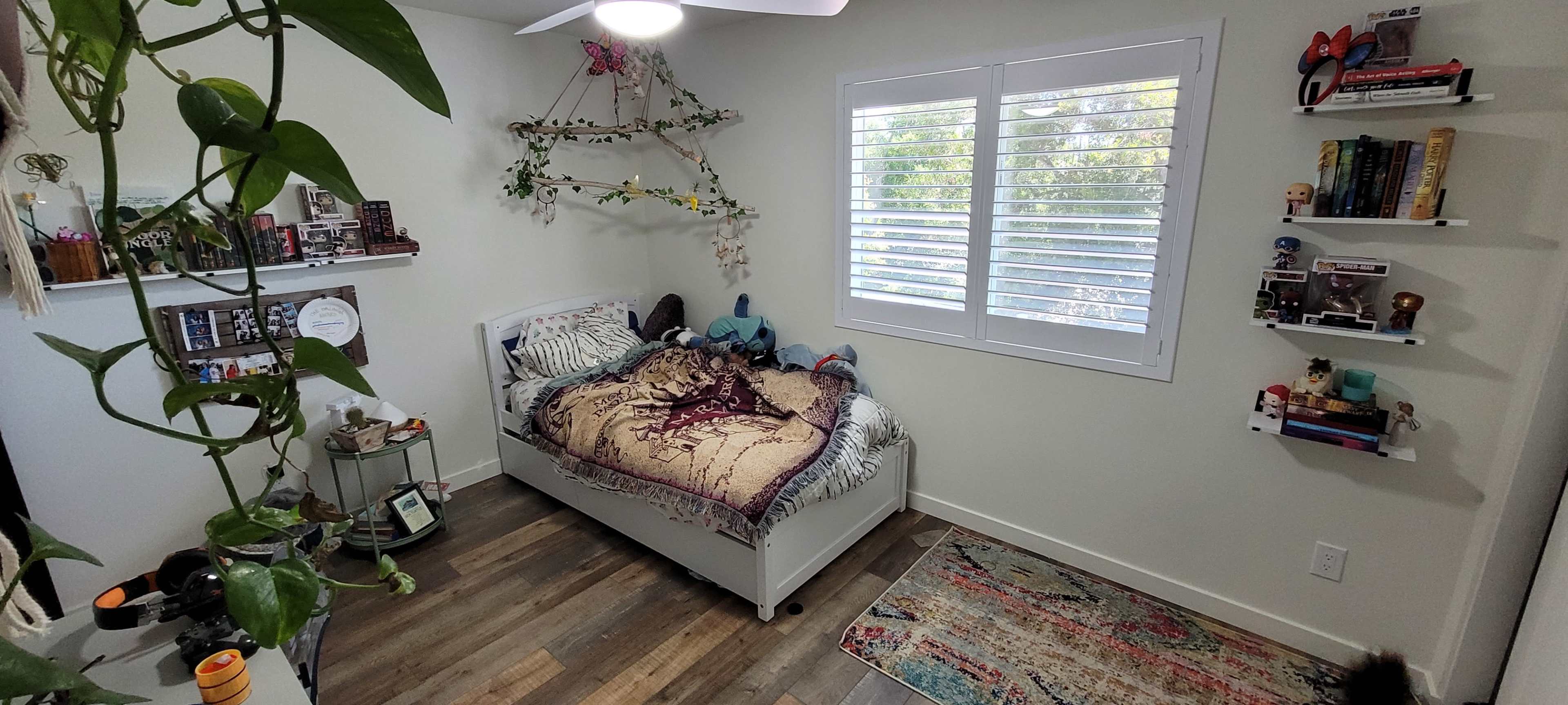 A small bedroom with a bed, decorative shelves on the walls, and a large window with white shutters.