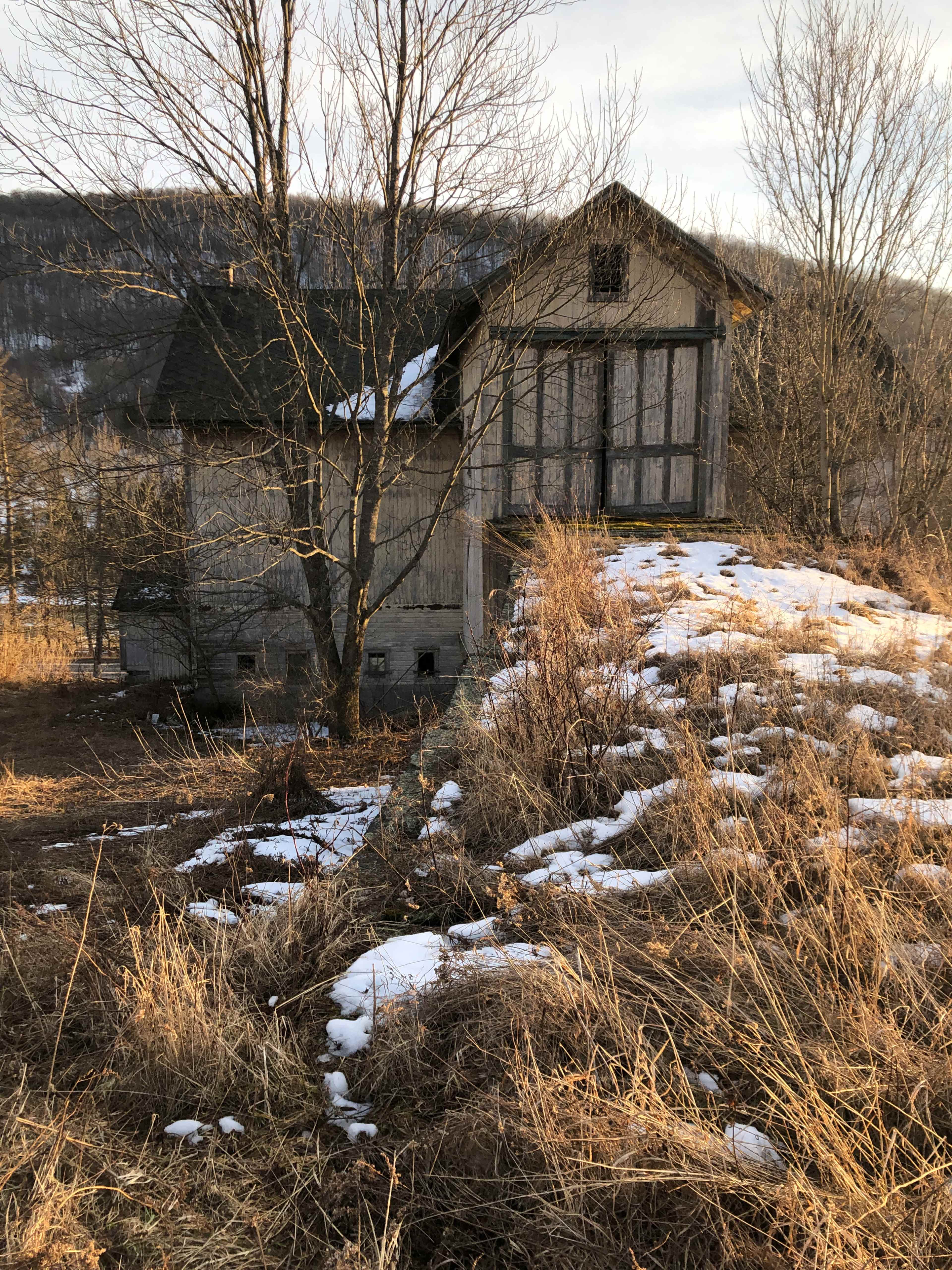 An abandoned wooden house, partially covered in snow, stands on a grassy hillside surrounded by bare trees in a rural landscape.
