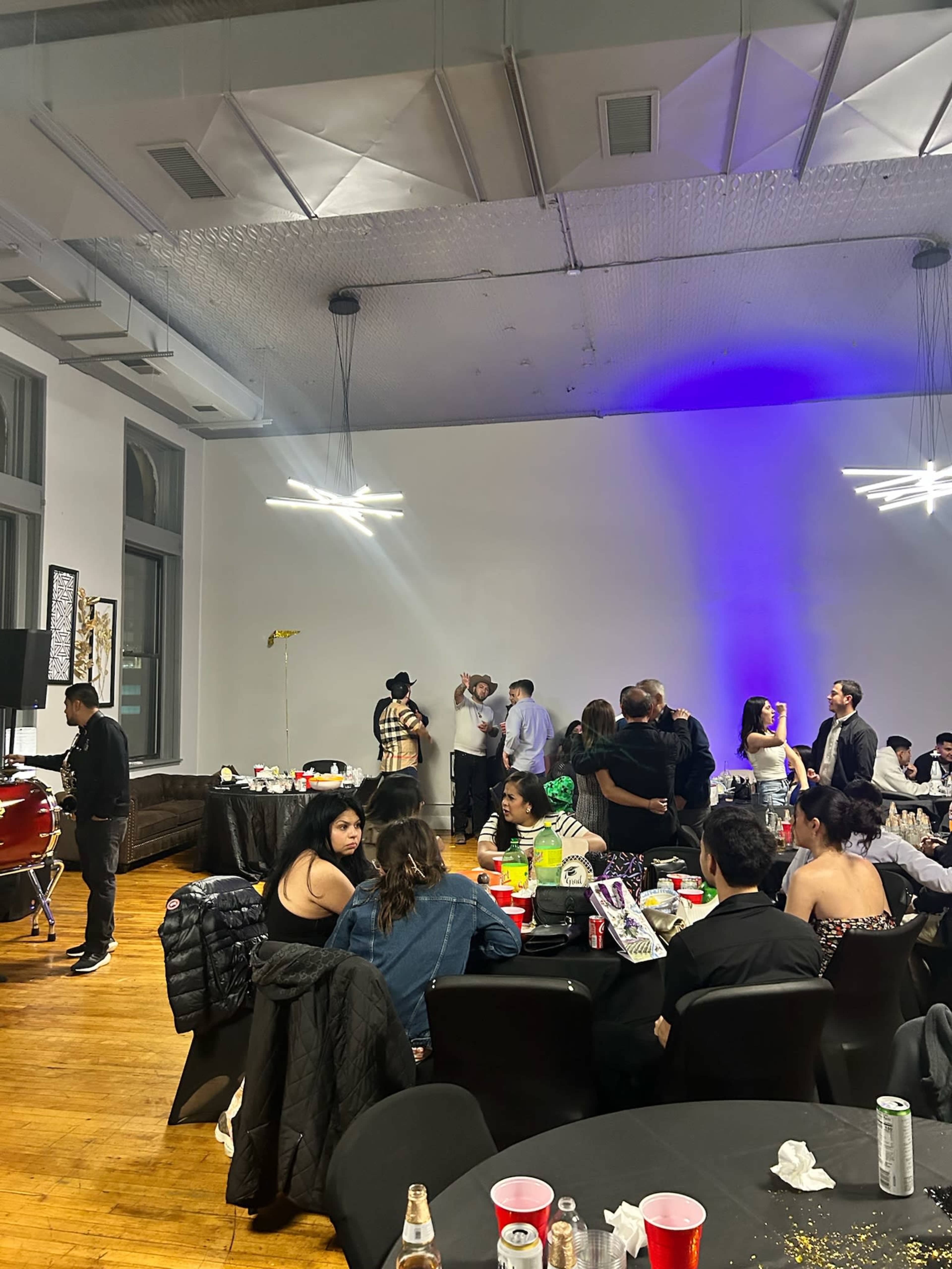 The image shows a crowded indoor event space with tables set for dining, people mingling, and decorations, including a projection of purple light on the wall.