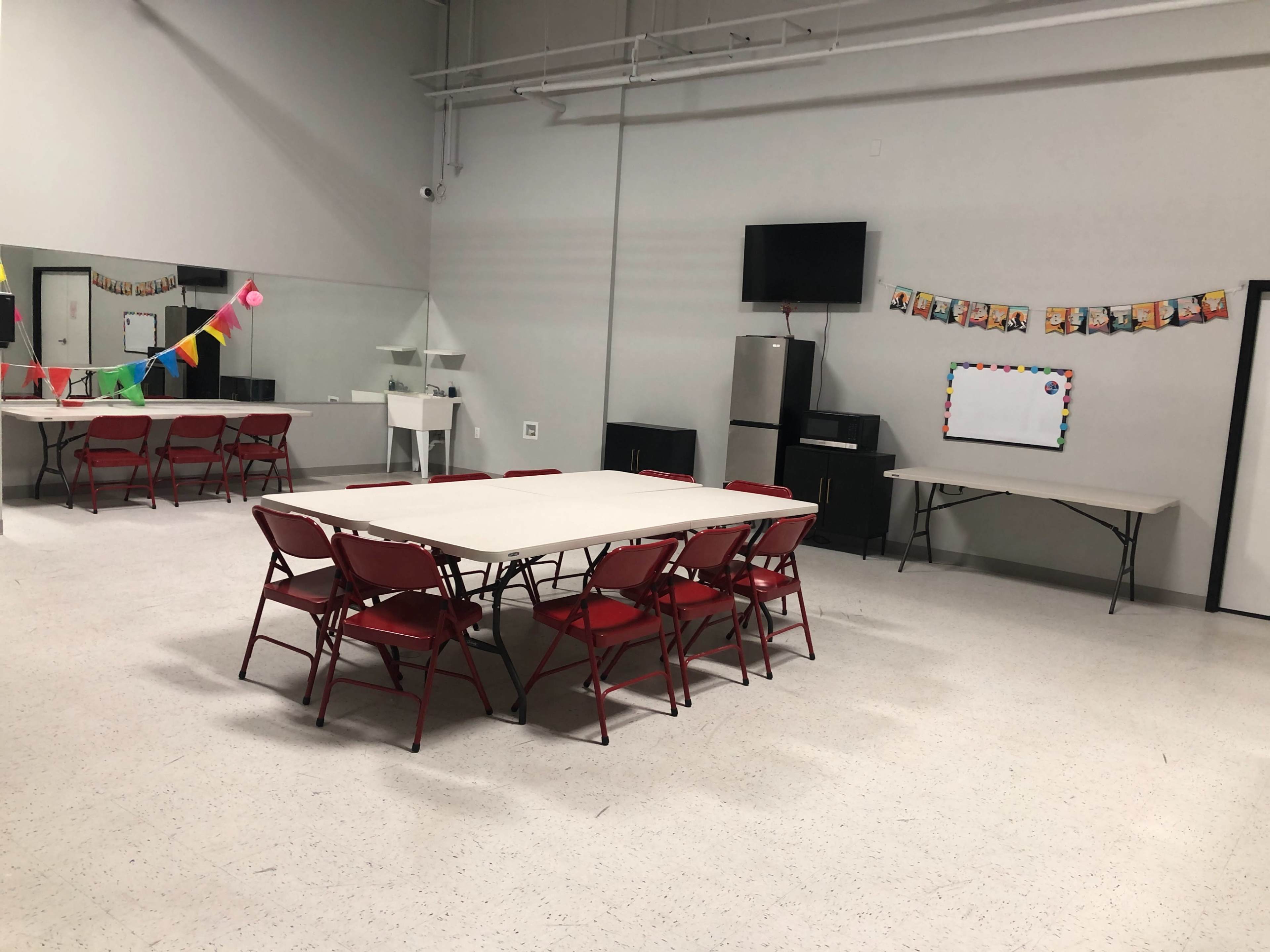 The image shows a spacious room with several tables and red folding chairs arranged, along with a mirrored wall and decorations hanging from the ceiling.
