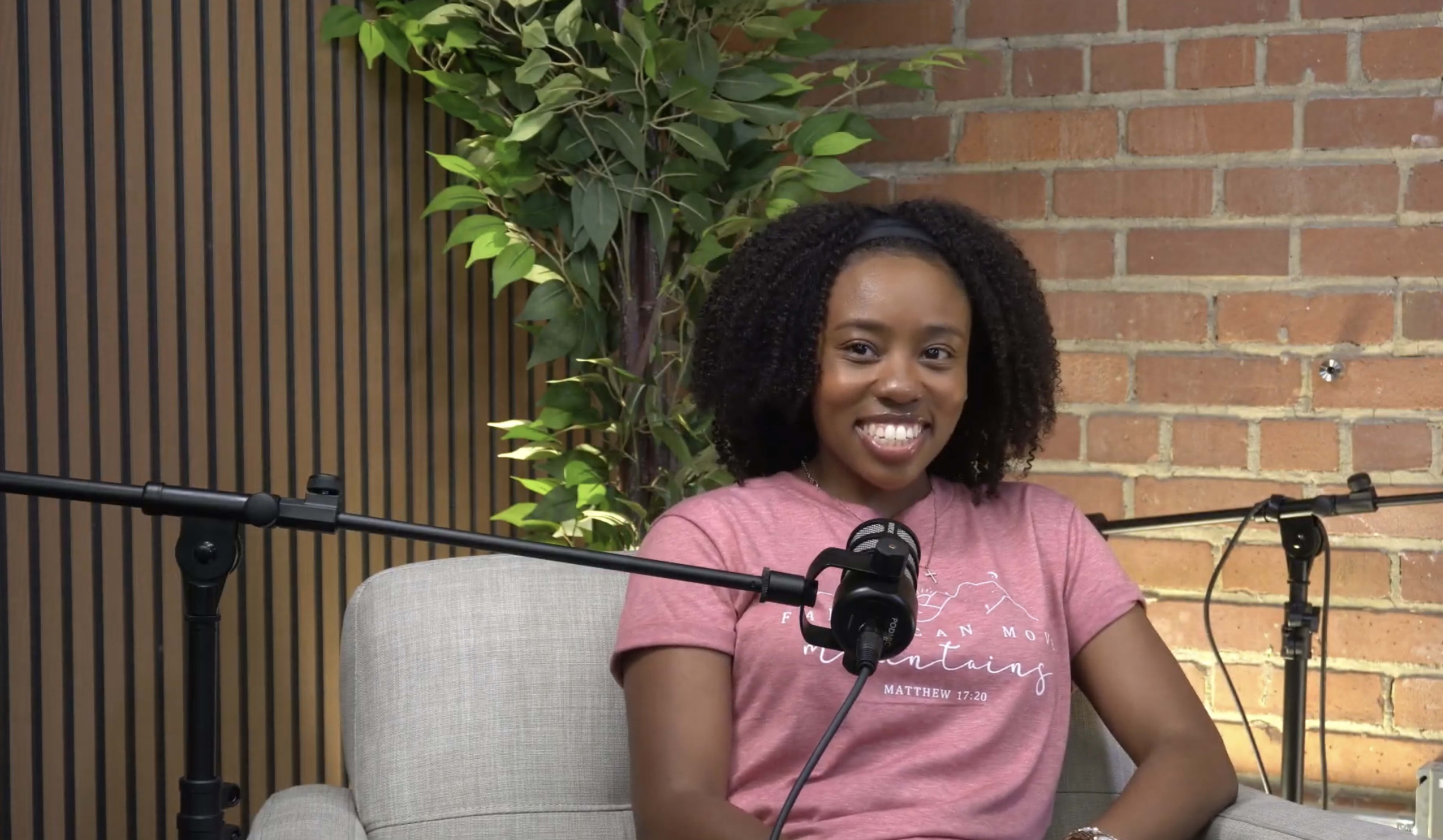 A woman with curly hair smiles while sitting on a sofa near a microphone and a plant against a brick wall.