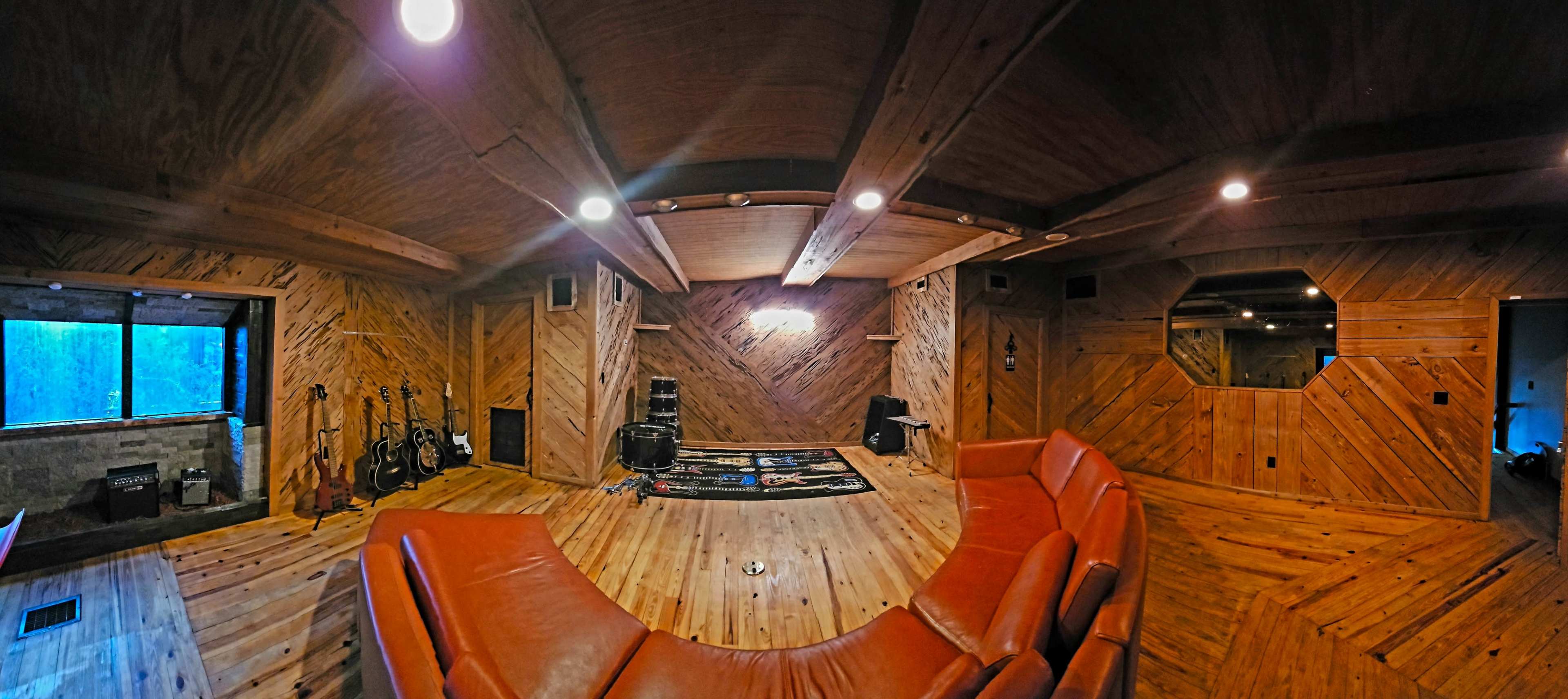 A spacious wooden-paneled room featuring a brown leather curved sofa, a patterned area rug, and musical instruments leaning against the walls.