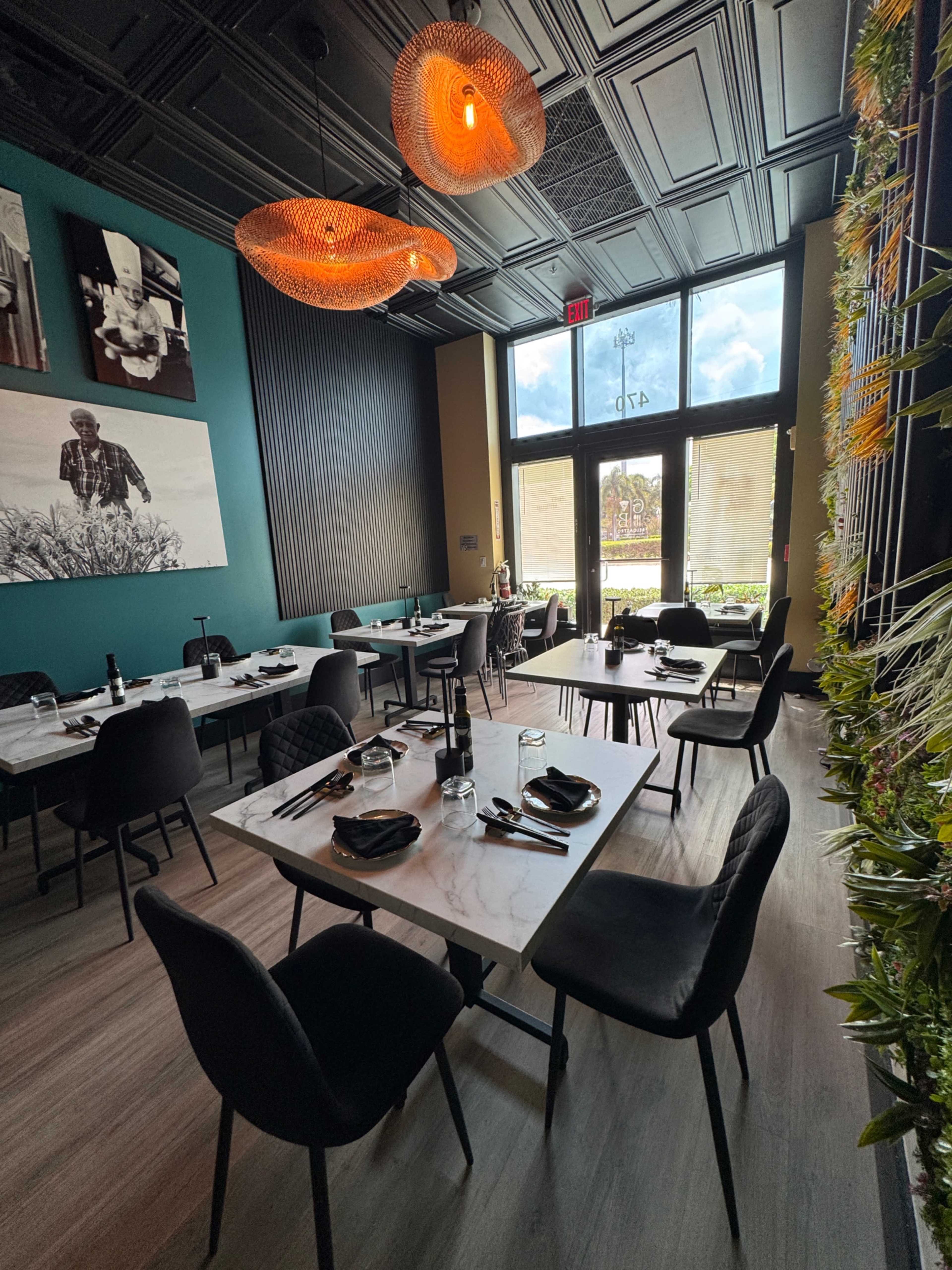 The image shows a modern restaurant interior featuring several tables set with black dinnerware, decorative light fixtures overhead, and large windows letting in natural light.