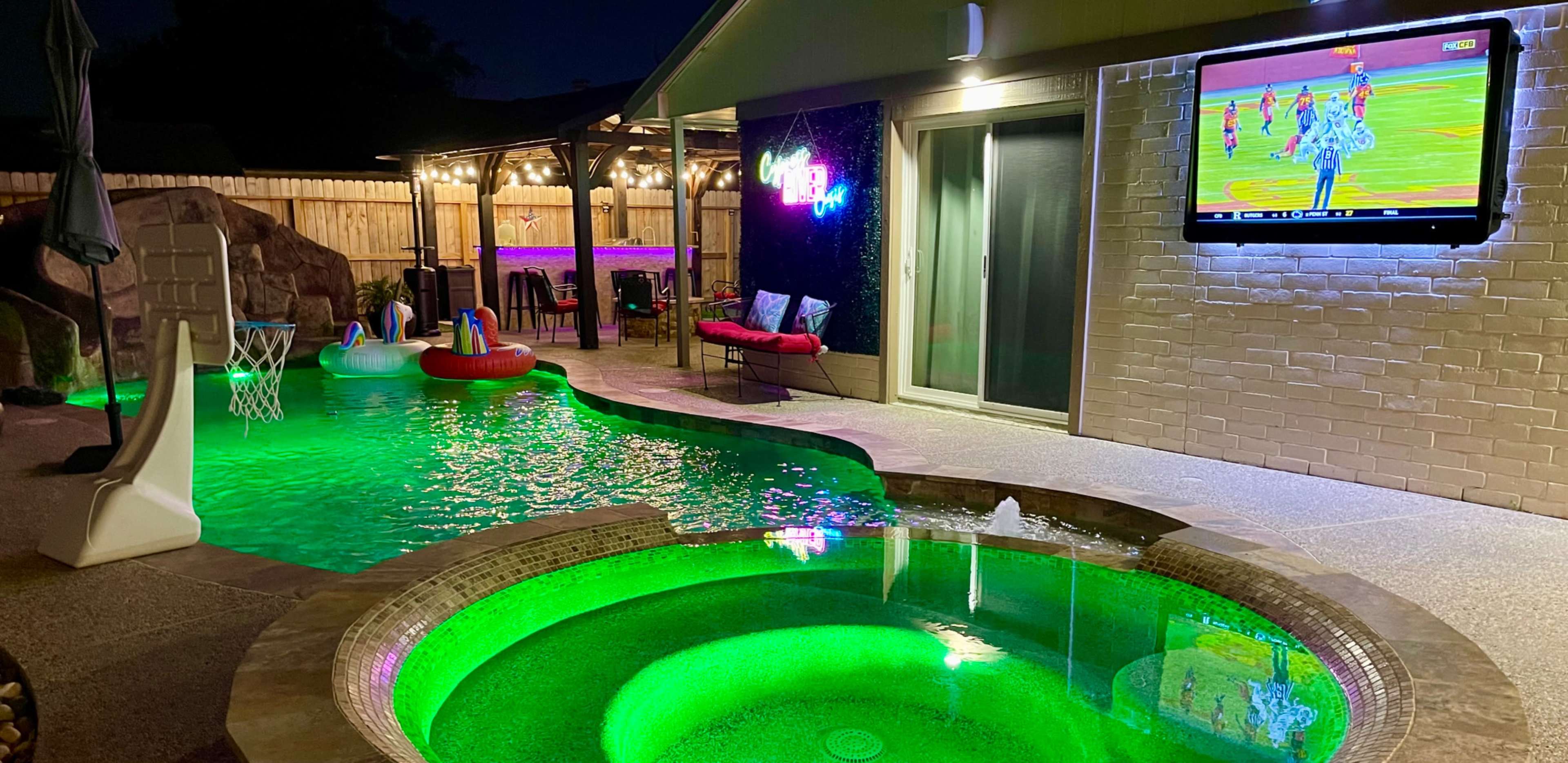 A backyard scene featuring a glowing pool with a spa, a TV mounted on a wall displaying a sports game, and seating areas illuminated by string lights.