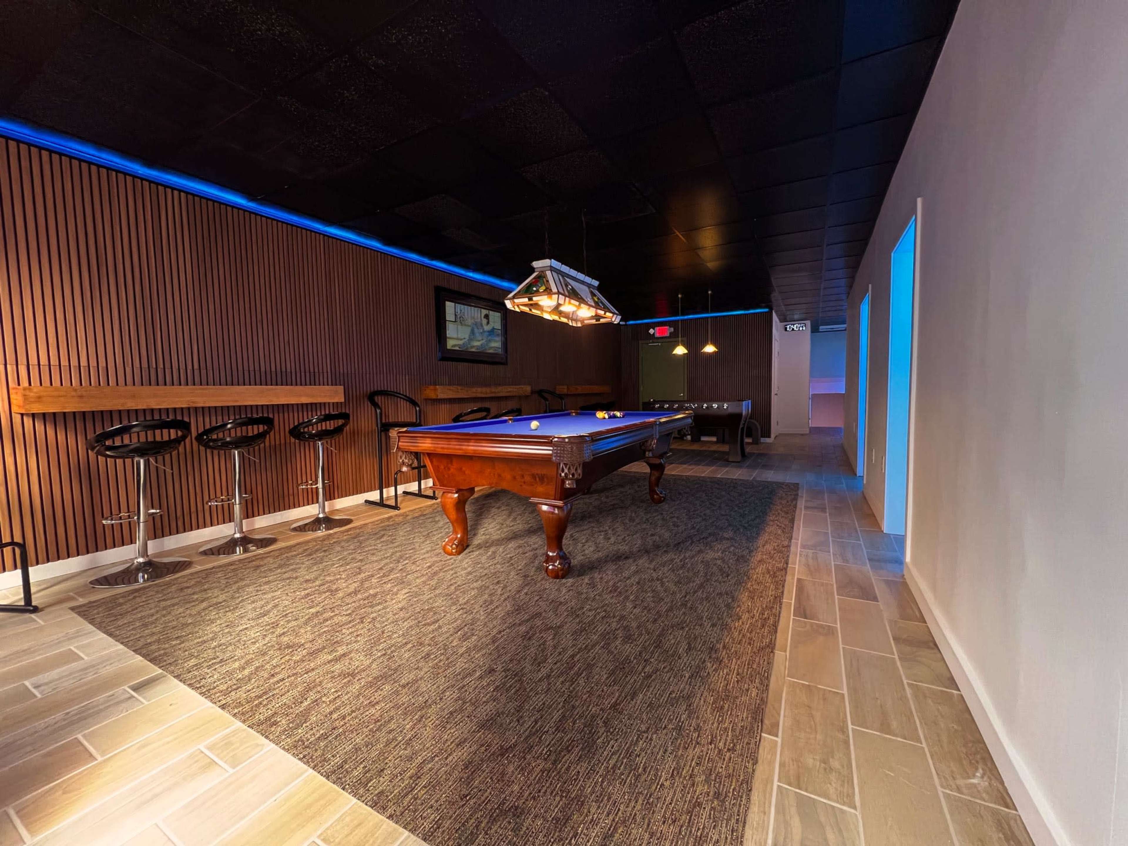 The image shows a modern game room featuring a pool table, bar seating, and ambient lighting along the walls.