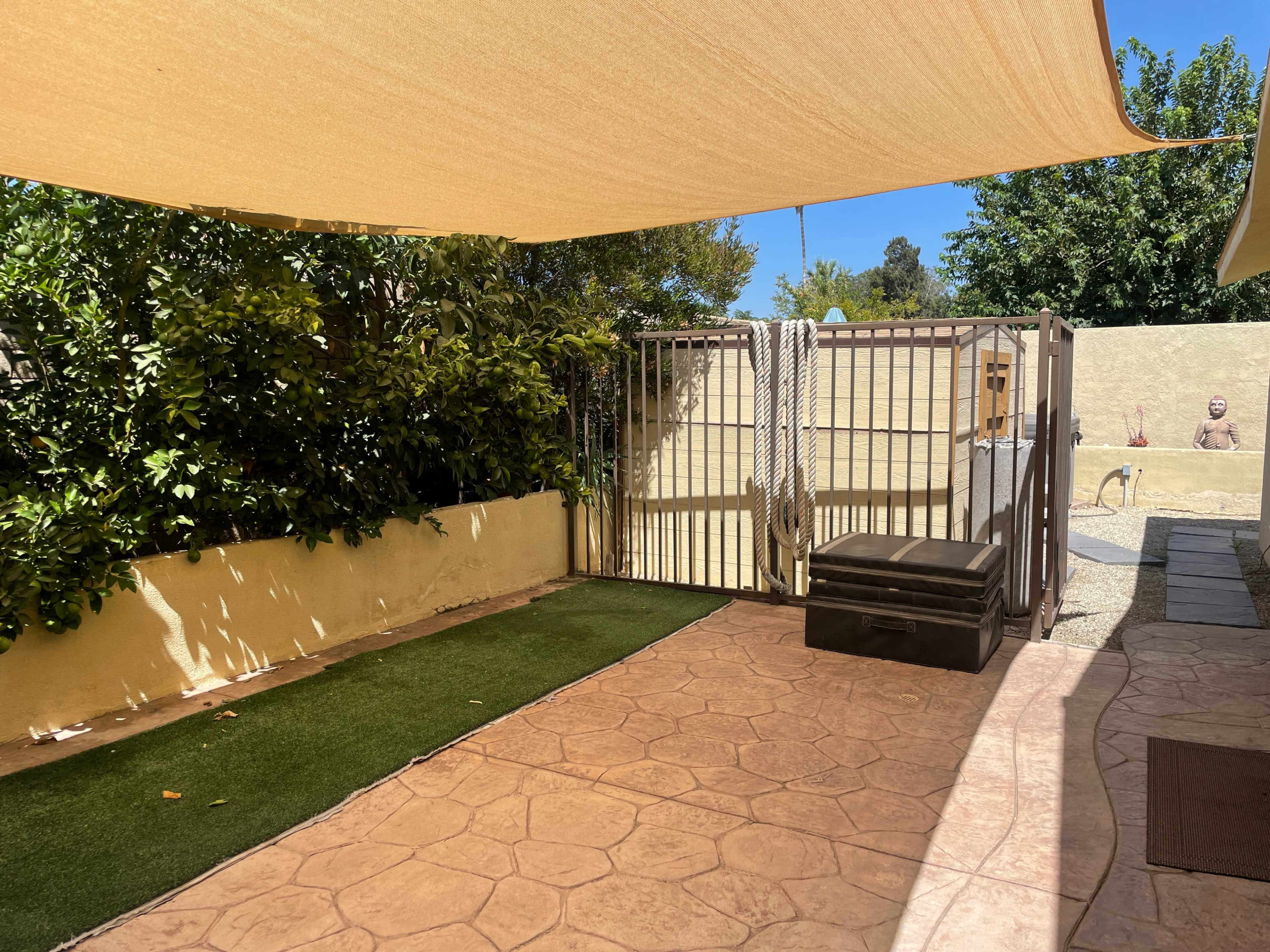 A patio area with a shaded section, artificial grass, a gated enclosure, and a storage box against a wall.