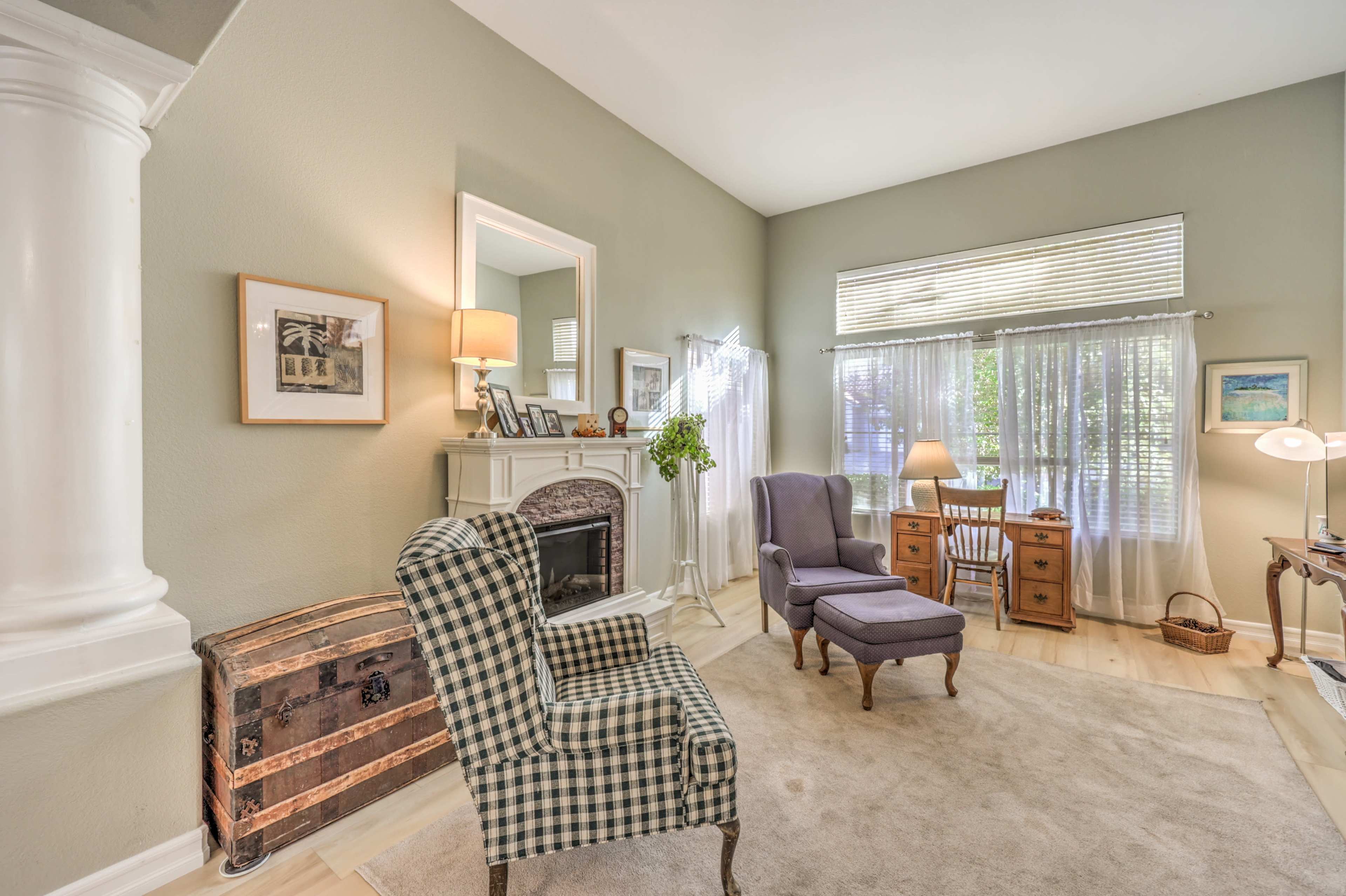 A cozy living room with a fireplace, two chairs, a small wooden table, and natural light coming through sheer curtains.