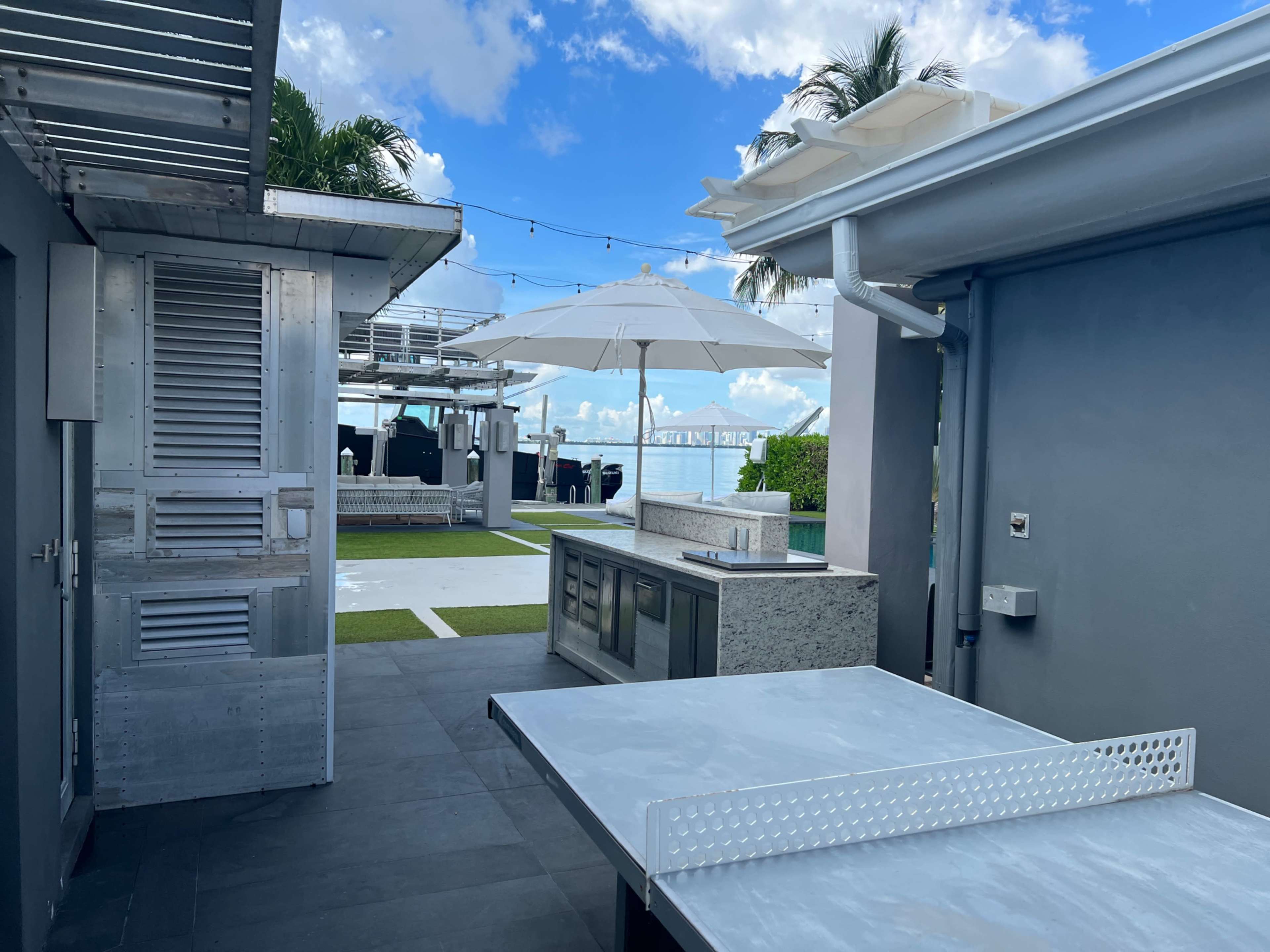 A modern outdoor kitchen area with a stainless steel countertop, an umbrella shading the seating area, and a view of a grassy space and palm trees.