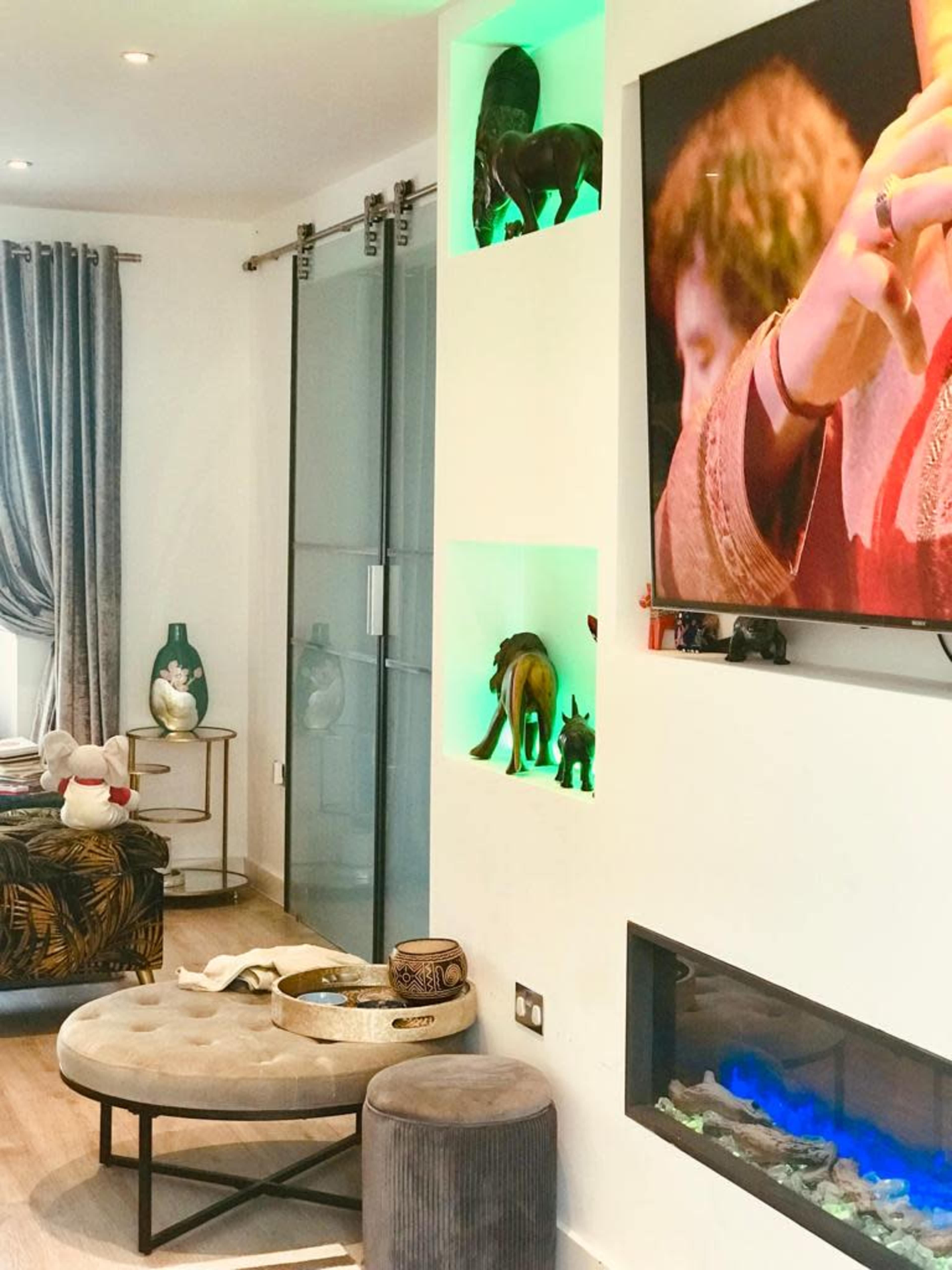 A modern living room featuring a seating area, decorative shelves with small sculptures, a large television on the wall, and an electric fireplace with blue lighting.
