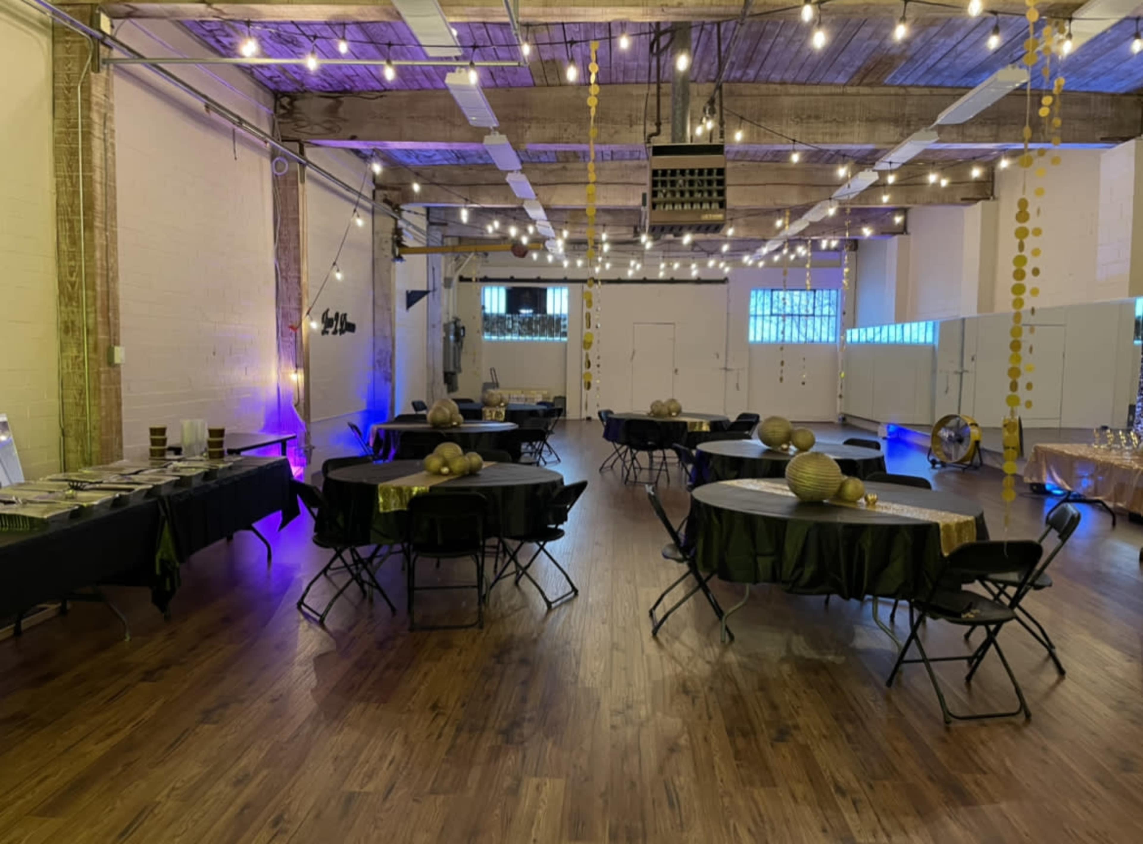 A spacious event hall set up with round tables covered in black and green tablecloths, illuminated by string lights and downlighting.