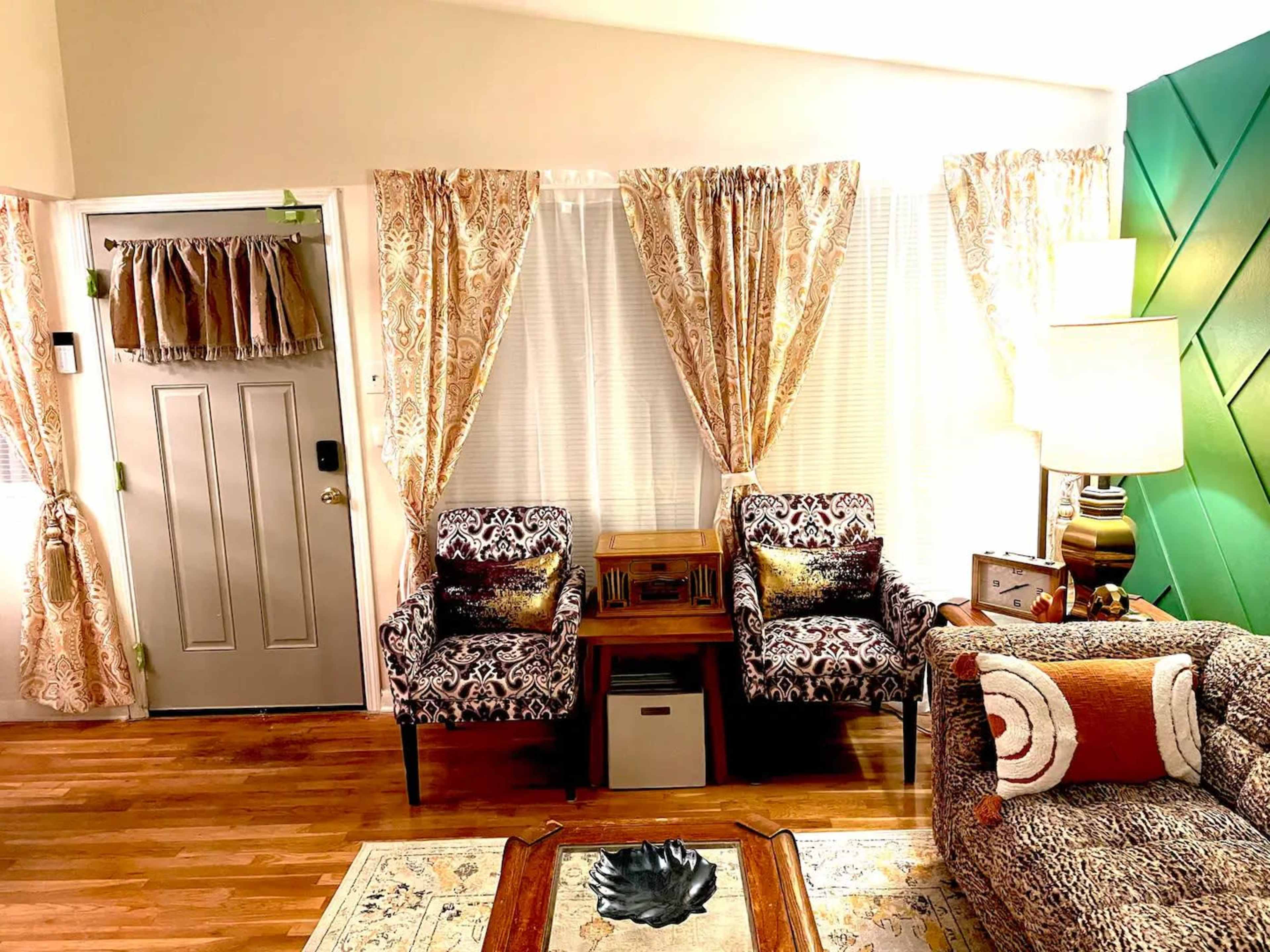 The room features two patterned armchairs, a sofa, a wooden coffee table with a decorative piece, and light-colored curtains at the windows.