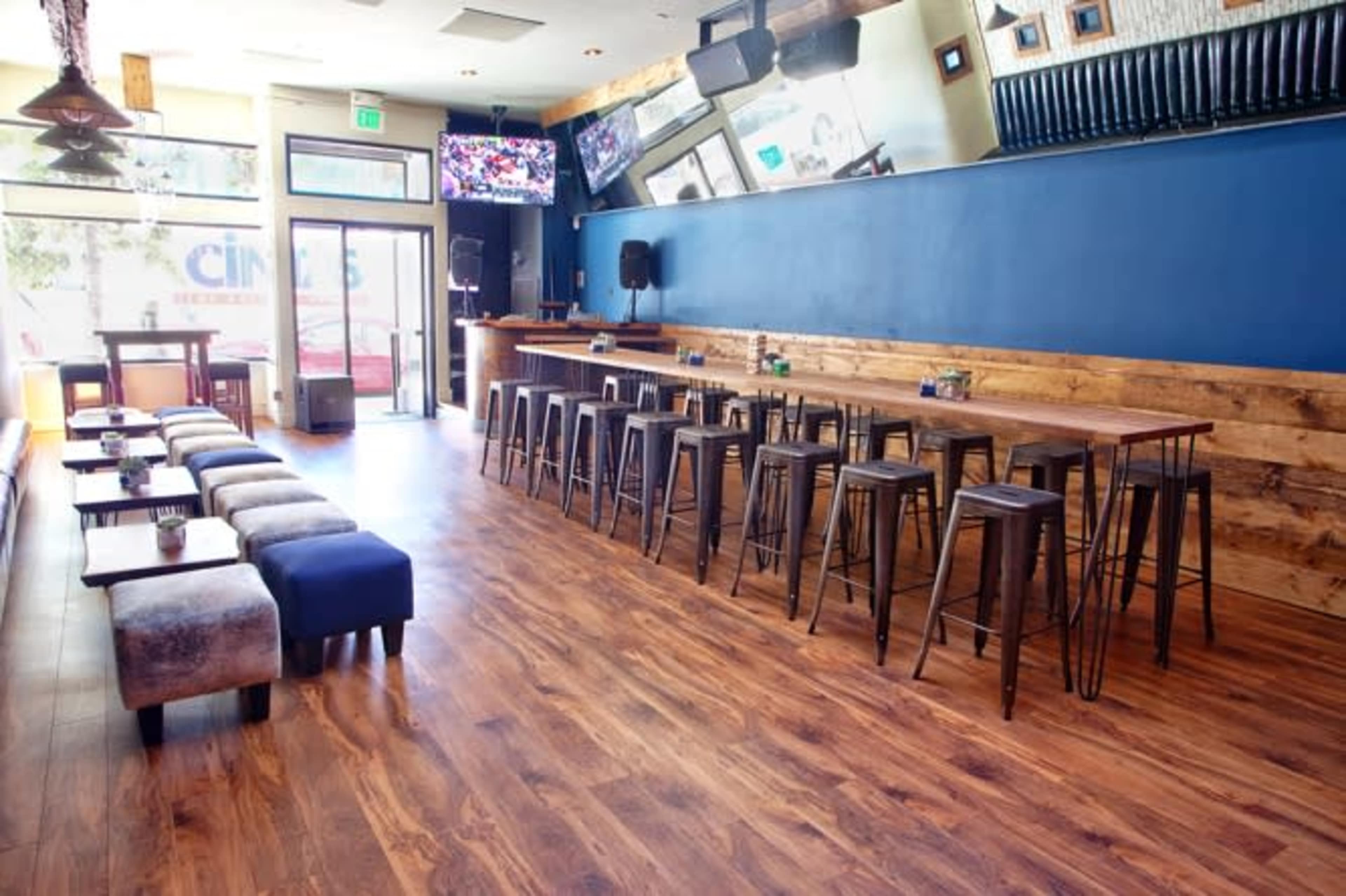 A modern bar interior with a long wooden table, high metal stools, and large windows allowing natural light.