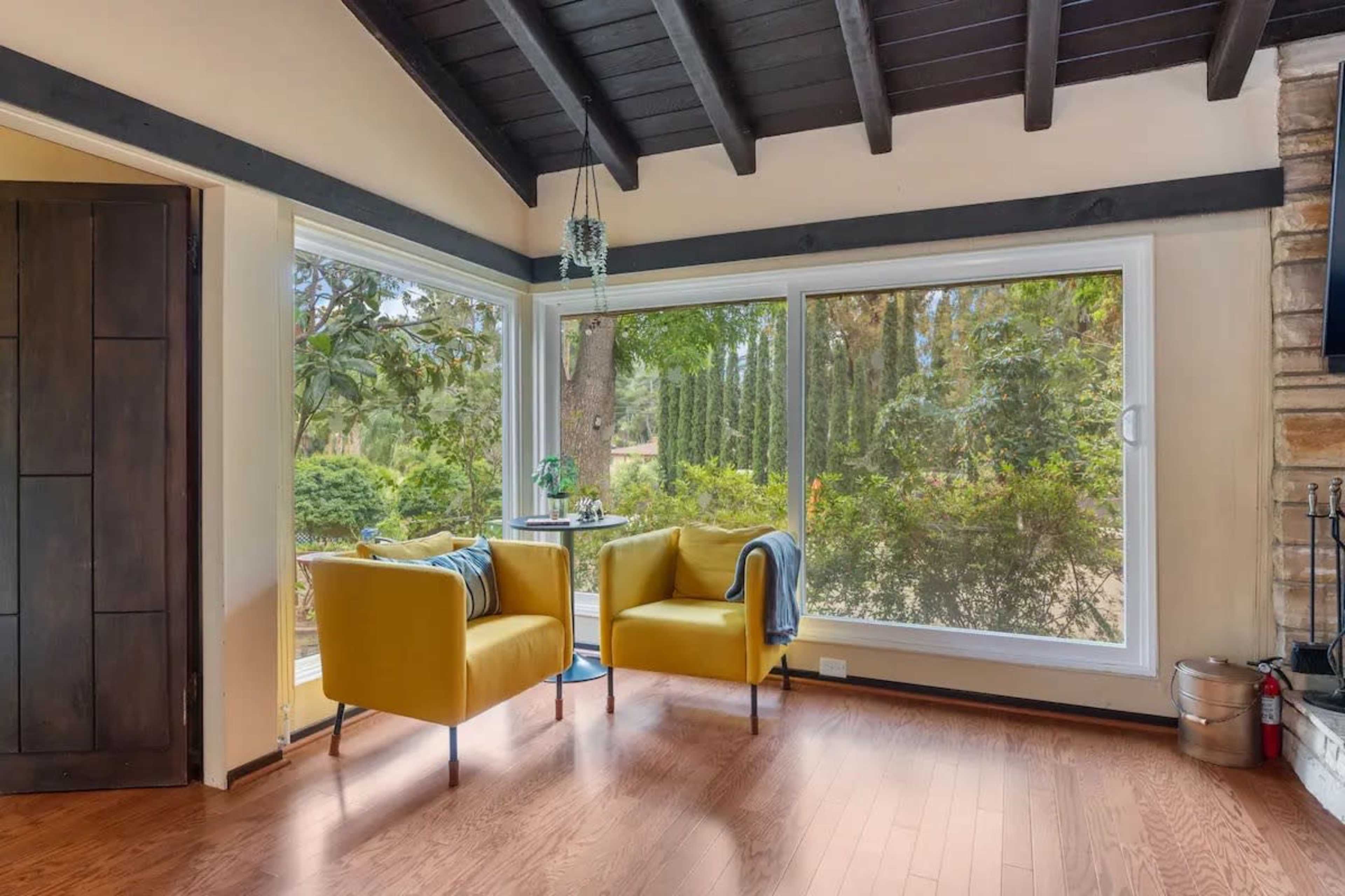 The image shows a sunlit corner of a room featuring two yellow chairs, large windows overlooking a garden, and wooden beams on the ceiling.