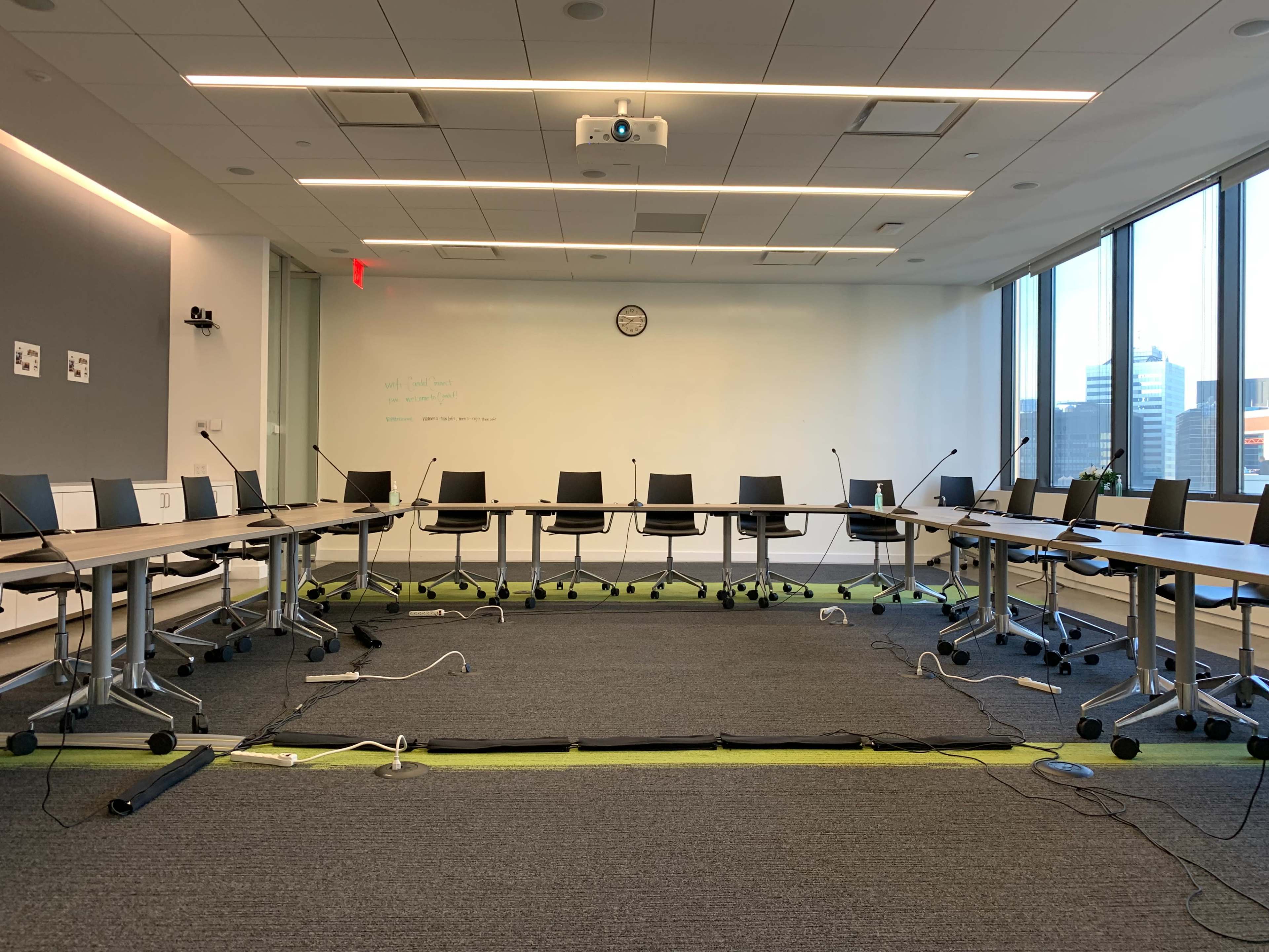 A conference room is set up with circular tables and black chairs arranged around them, featuring a projector and several microphones, with large windows providing natural light.