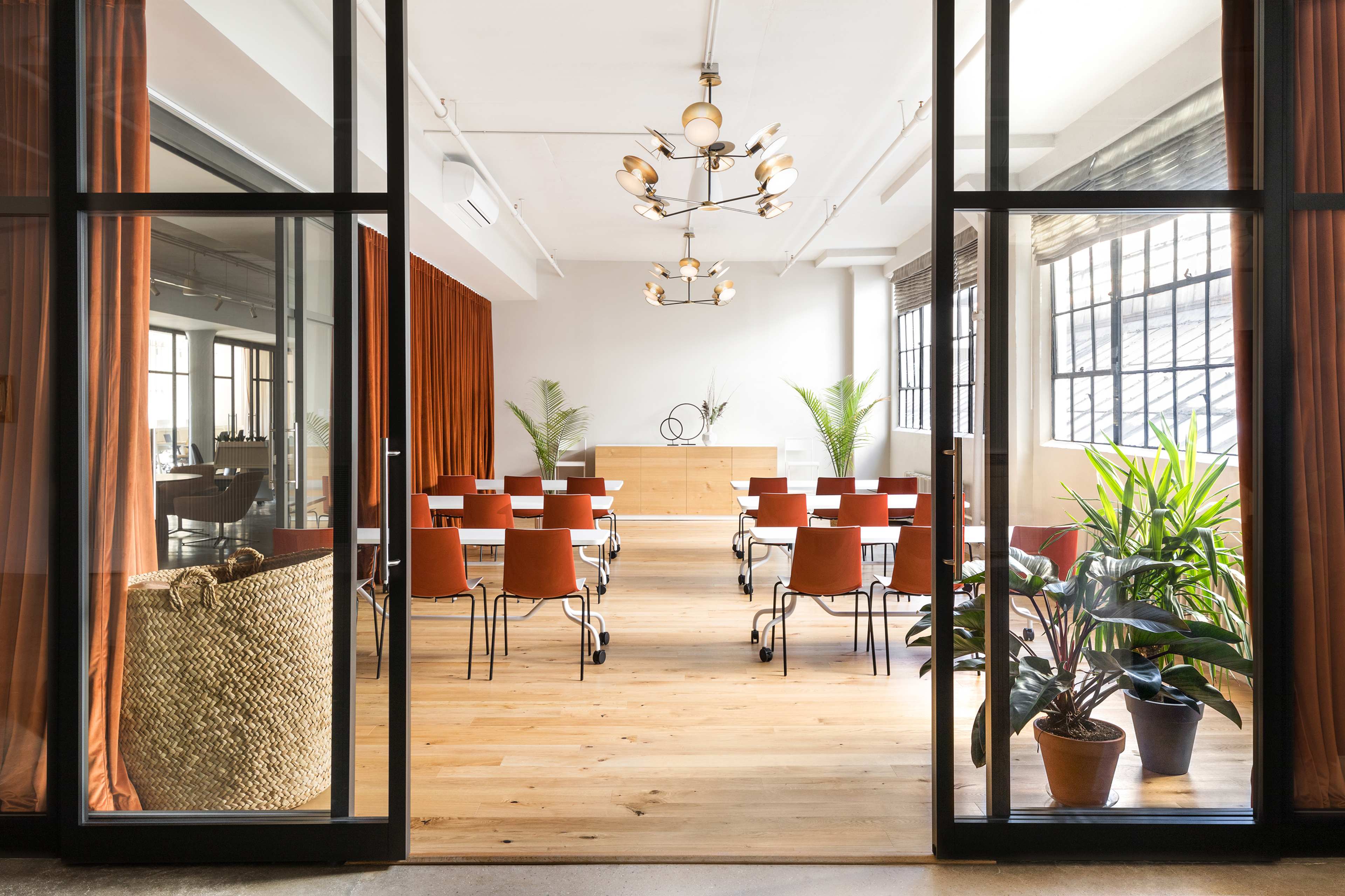 A spacious meeting room features rows of chairs facing a wooden wall, framed by large windows and surrounded by plants.