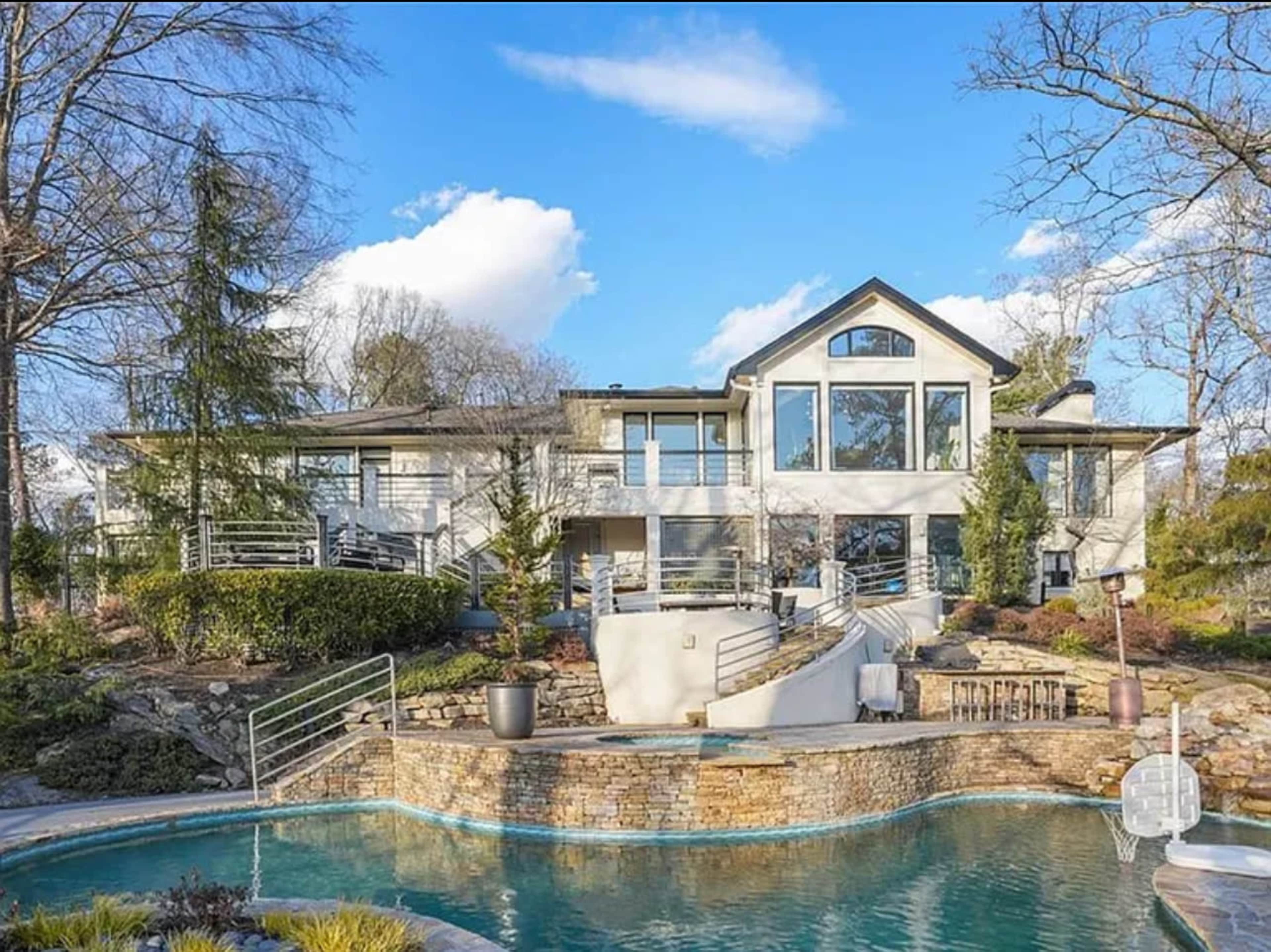 A large modern house with multiple windows is situated next to a swimming pool and surrounded by landscaped gardens and trees.