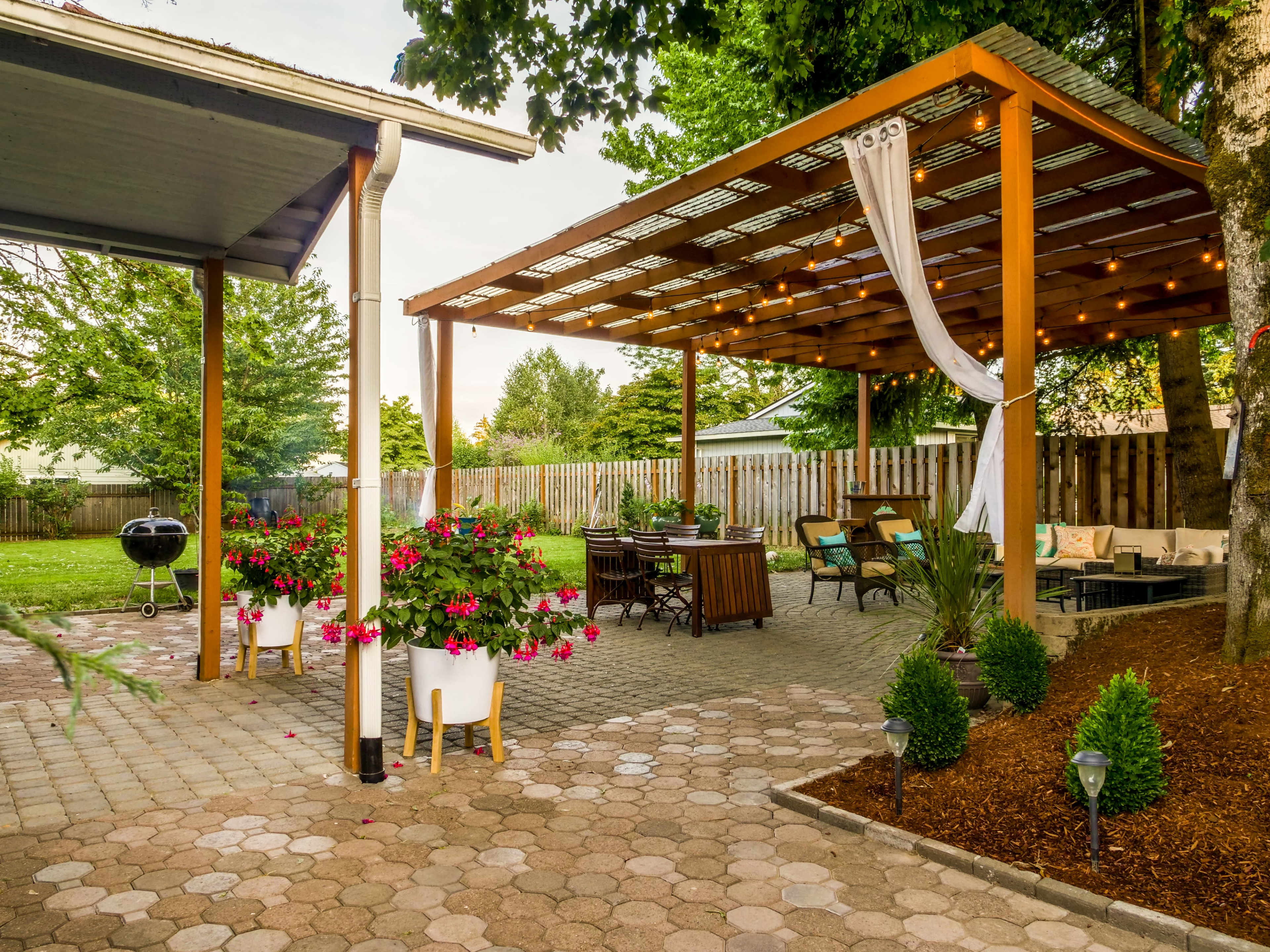 A backyard patio with a wooden pergola, outdoor seating, hanging lights, and flower pots next to a paved walkway.