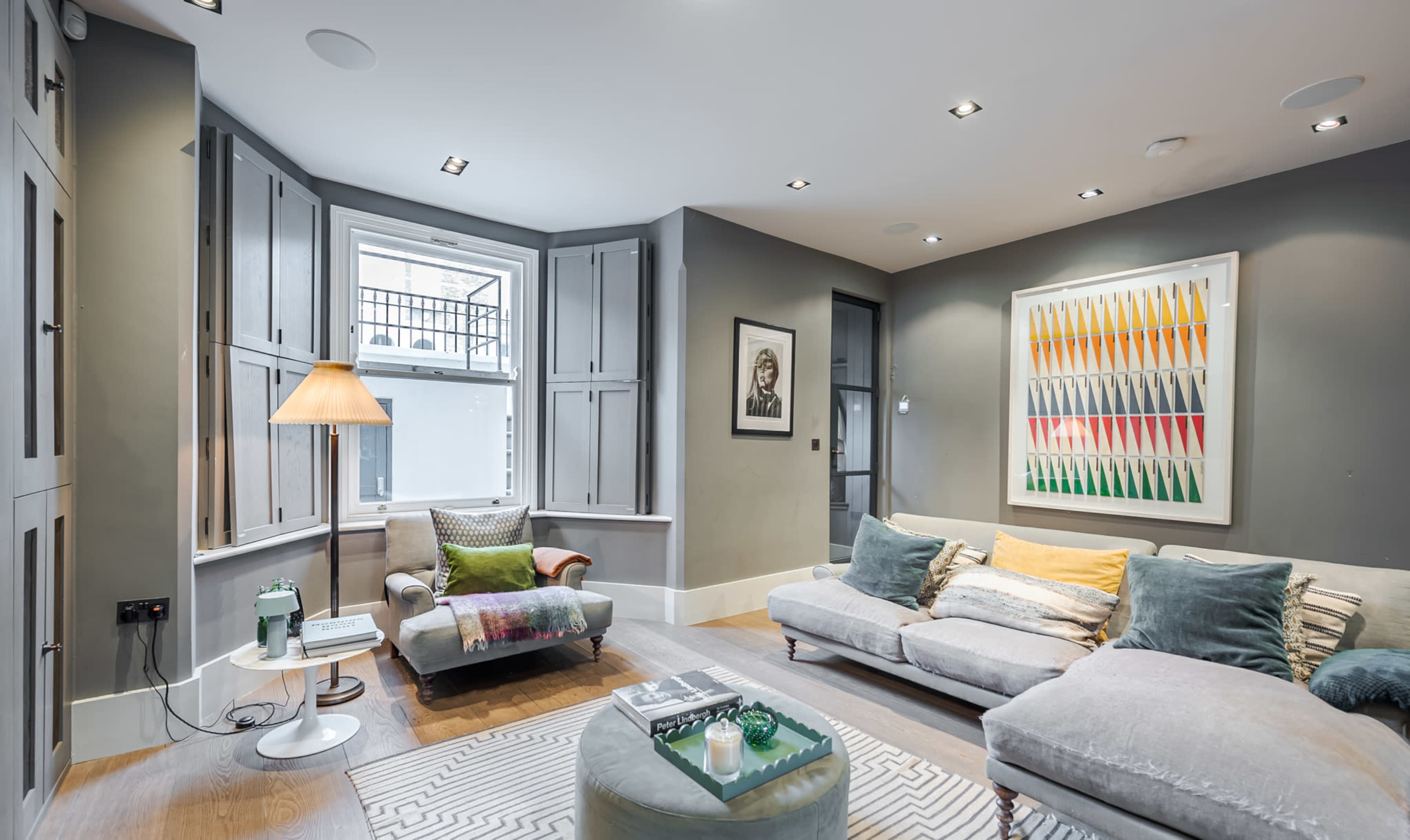 A modern living room features a sectional sofa, a patterned rug, and bay windows with closed shutters.