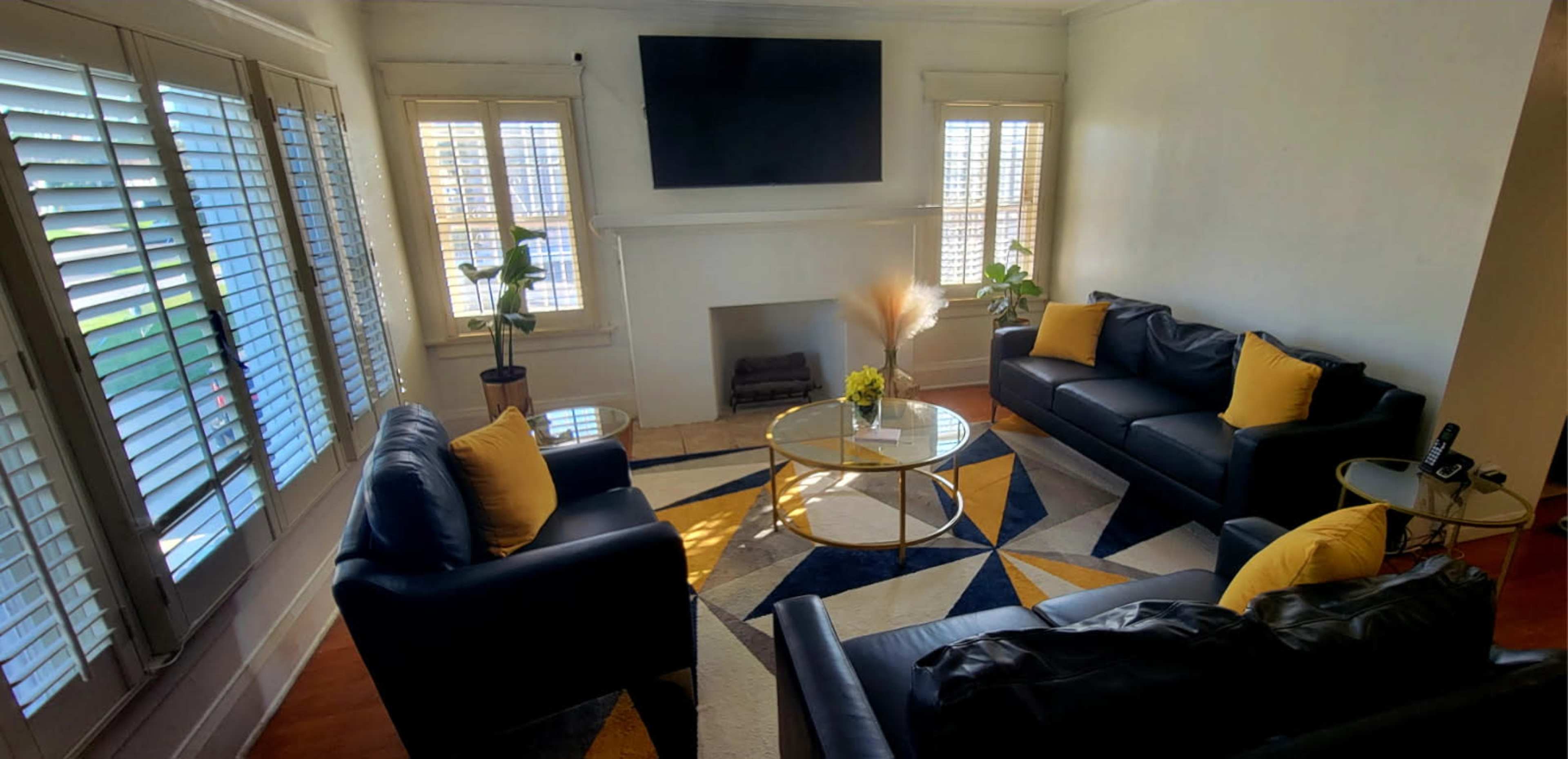The image shows a modern living room with black leather sofas, a circular glass coffee table, a flat-screen television mounted on the wall, and natural light coming through window shutters.