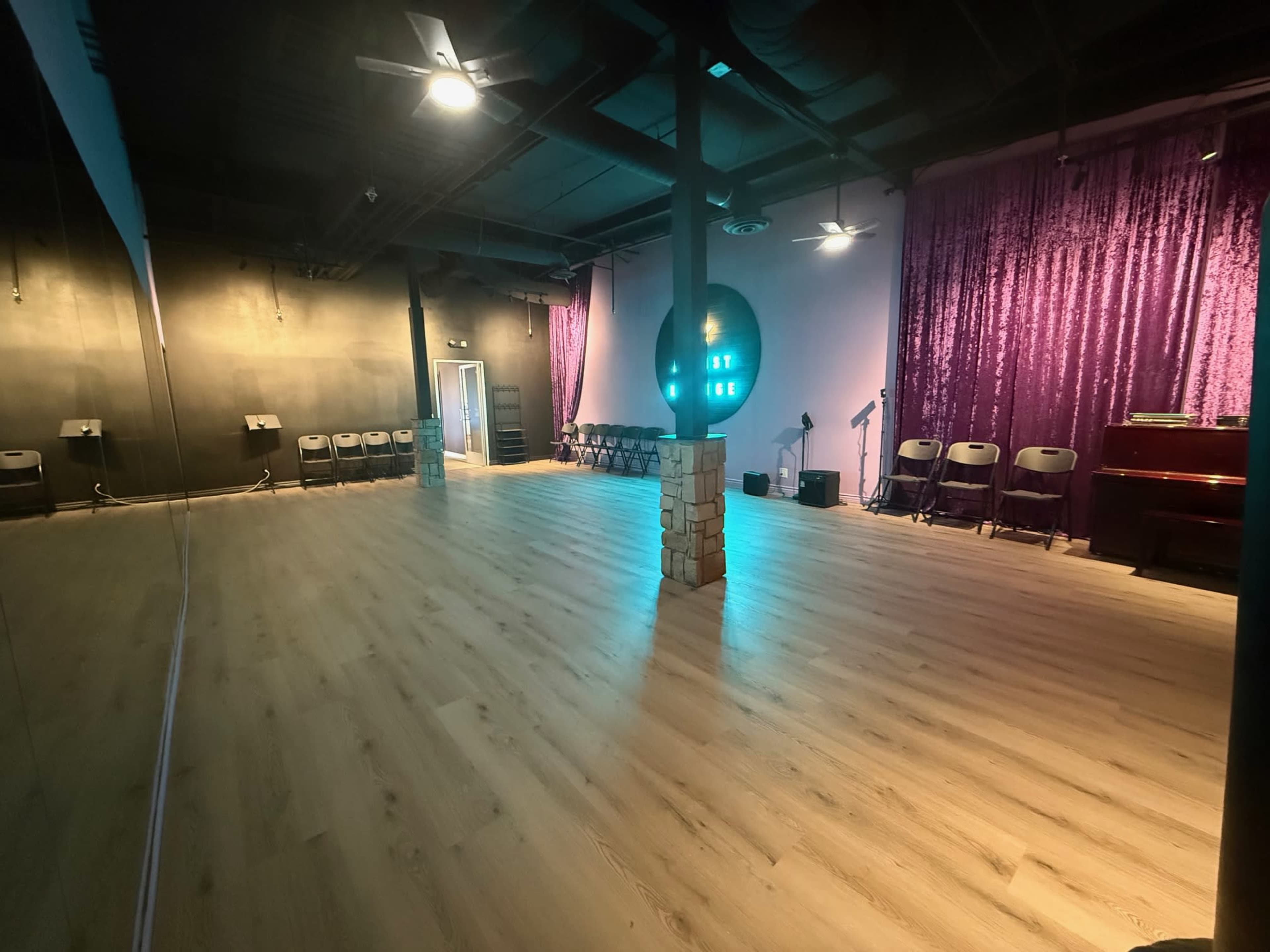 The image shows a spacious room with wooden flooring, purple curtains along one wall, a microphone stand, and chairs arranged in a semi-circle.