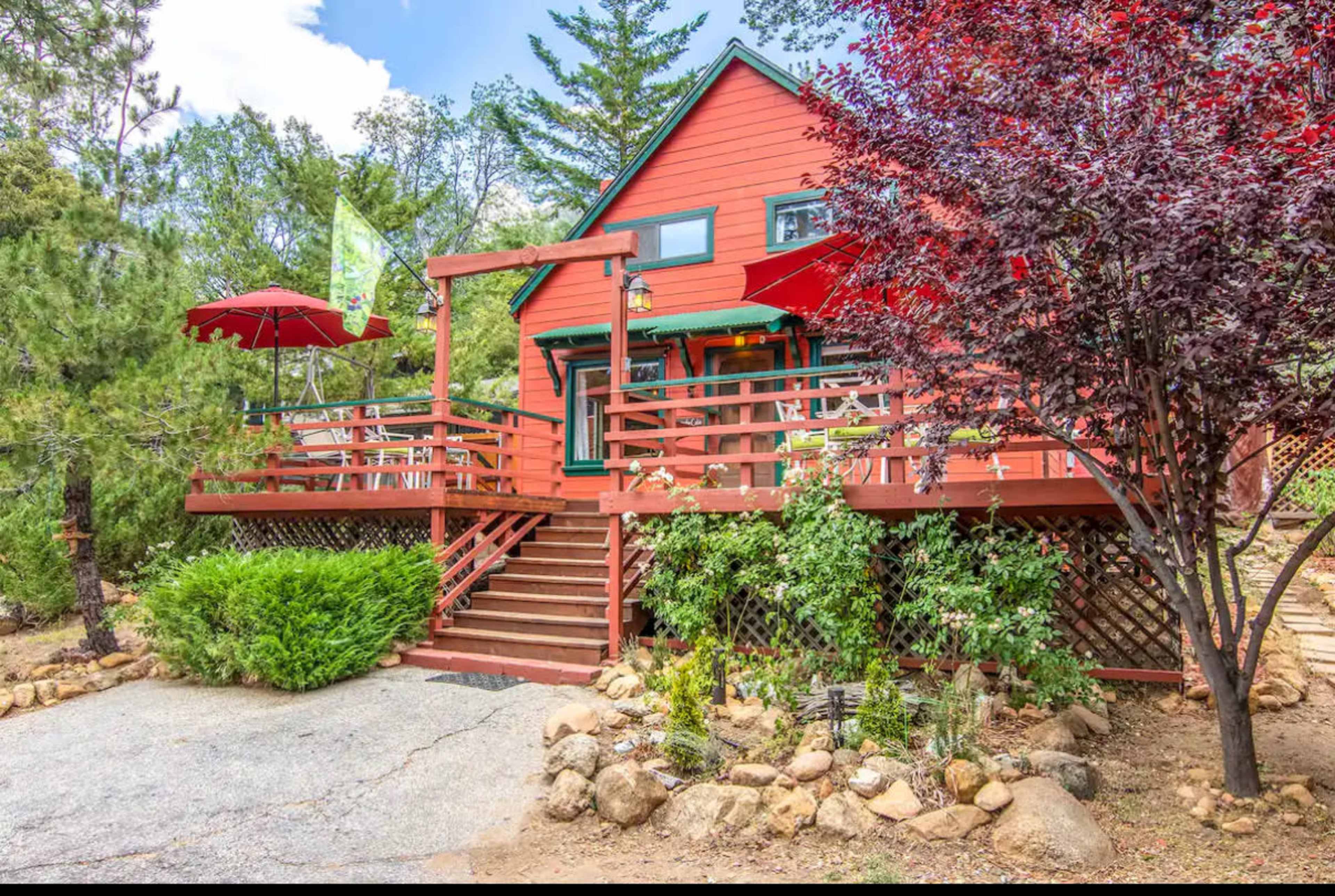 A red wooden cabin with a wraparound deck and outdoor seating is surrounded by trees and landscaped gardens.