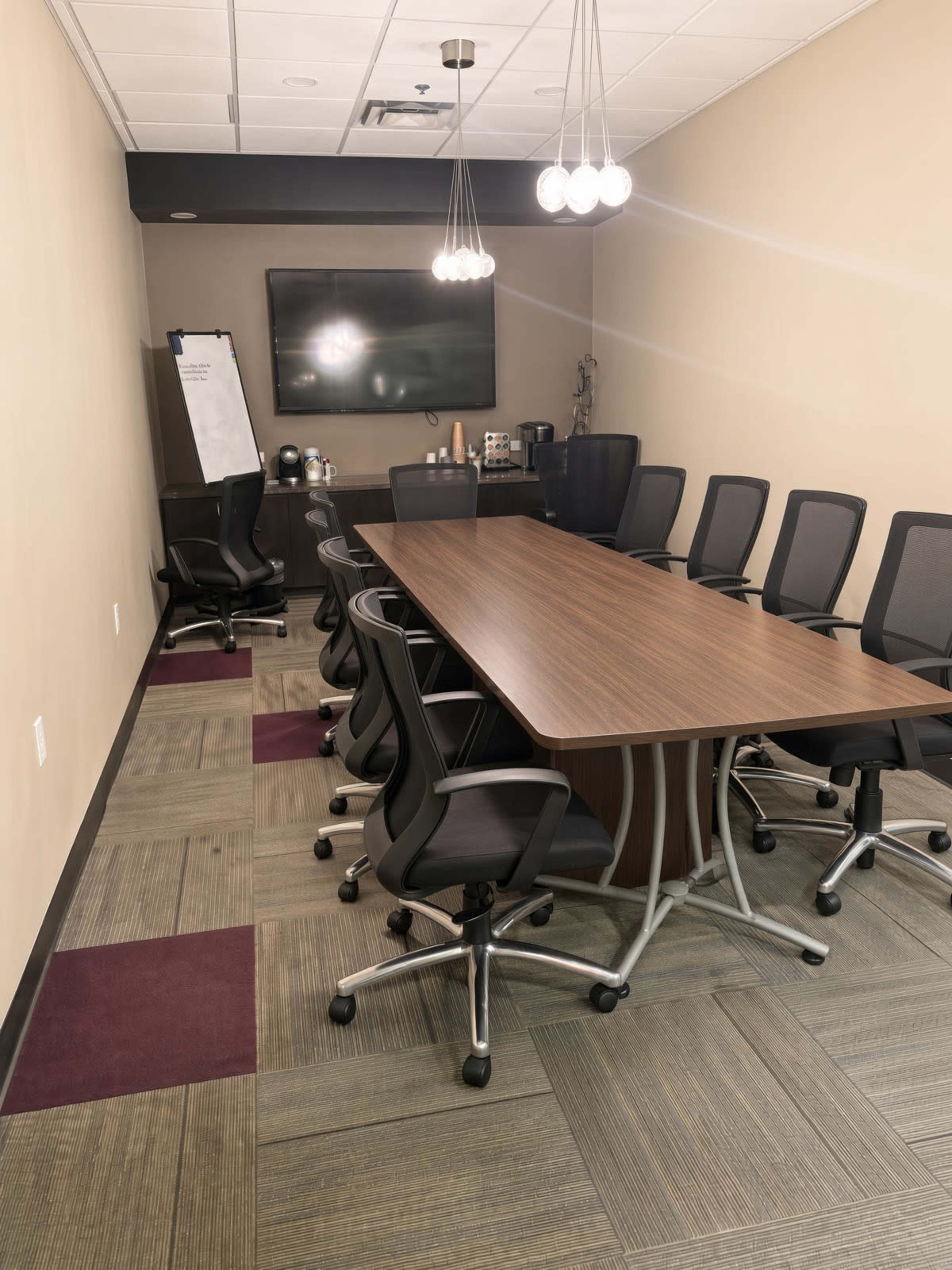 The image shows a modern conference room with a long wooden table, ergonomic chairs, a television mounted on the wall, and a whiteboard in the corner.