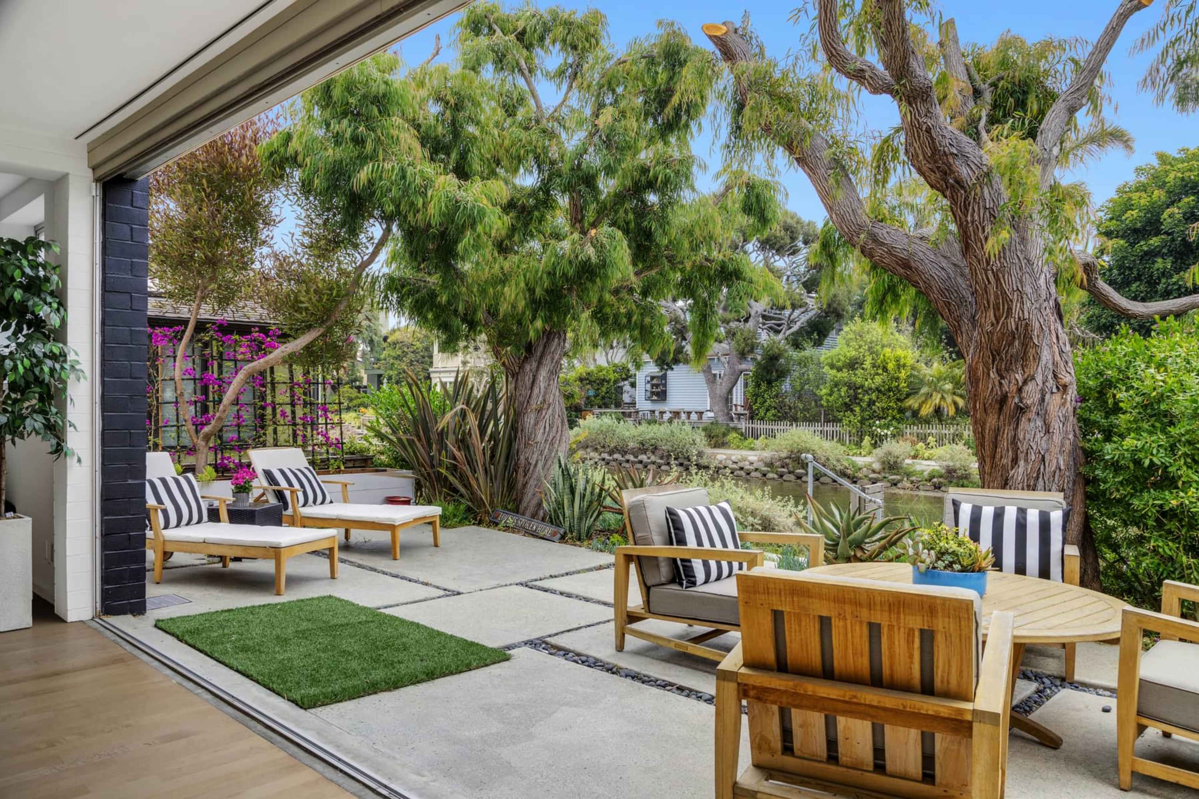 A modern patio features lounge chairs and a wooden table surrounded by greenery and trees.