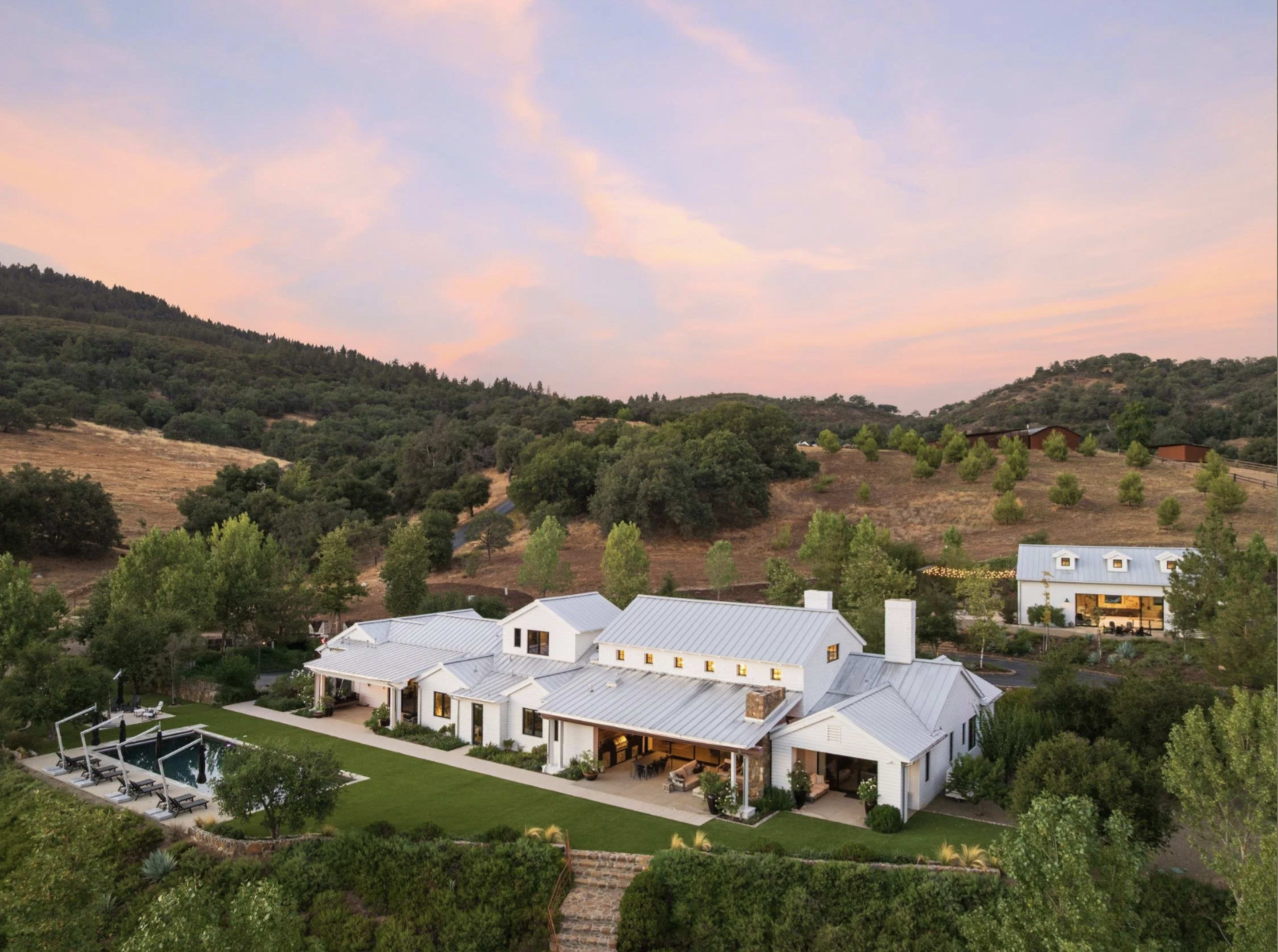A spacious white farmhouse with a metal roof is set against a hillside, surrounded by greenery and a swimming pool.