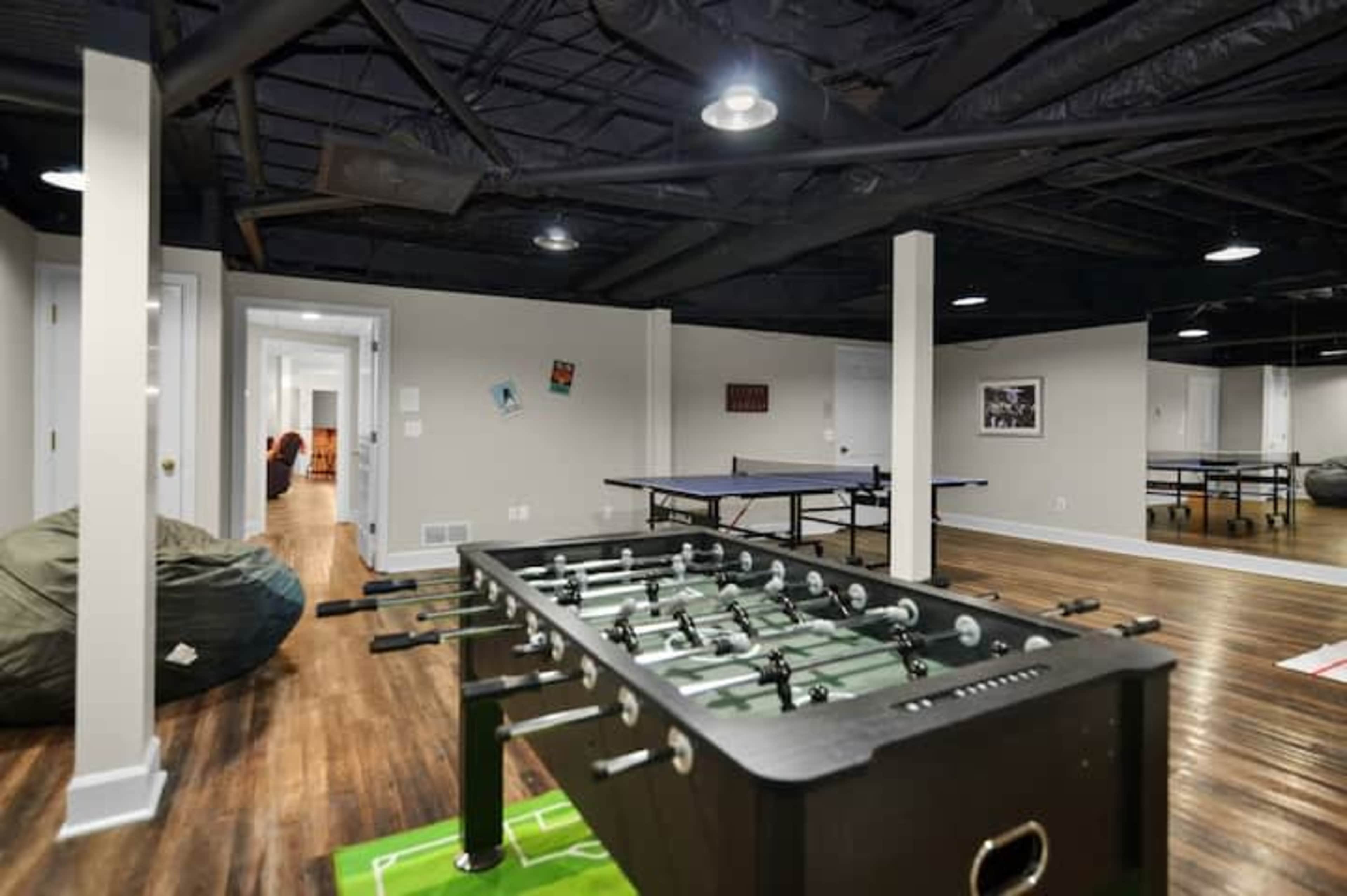 The image shows a spacious game room featuring a foosball table, ping pong table, and bean bag chairs, with a hallway leading to another room in the background.