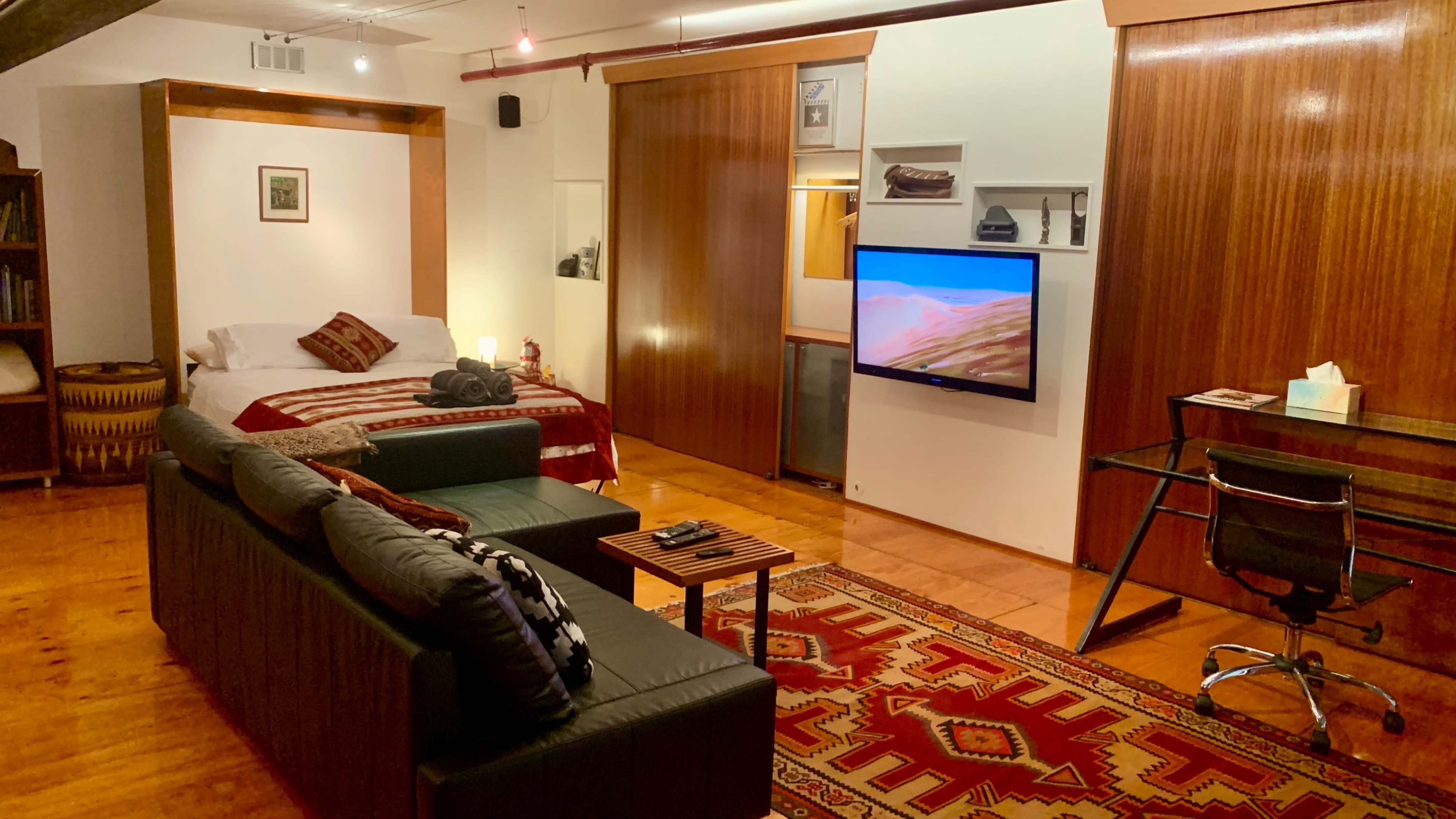 A modern studio apartment featuring a bed, a living area with a sofa, a wall-mounted television, and a desk.