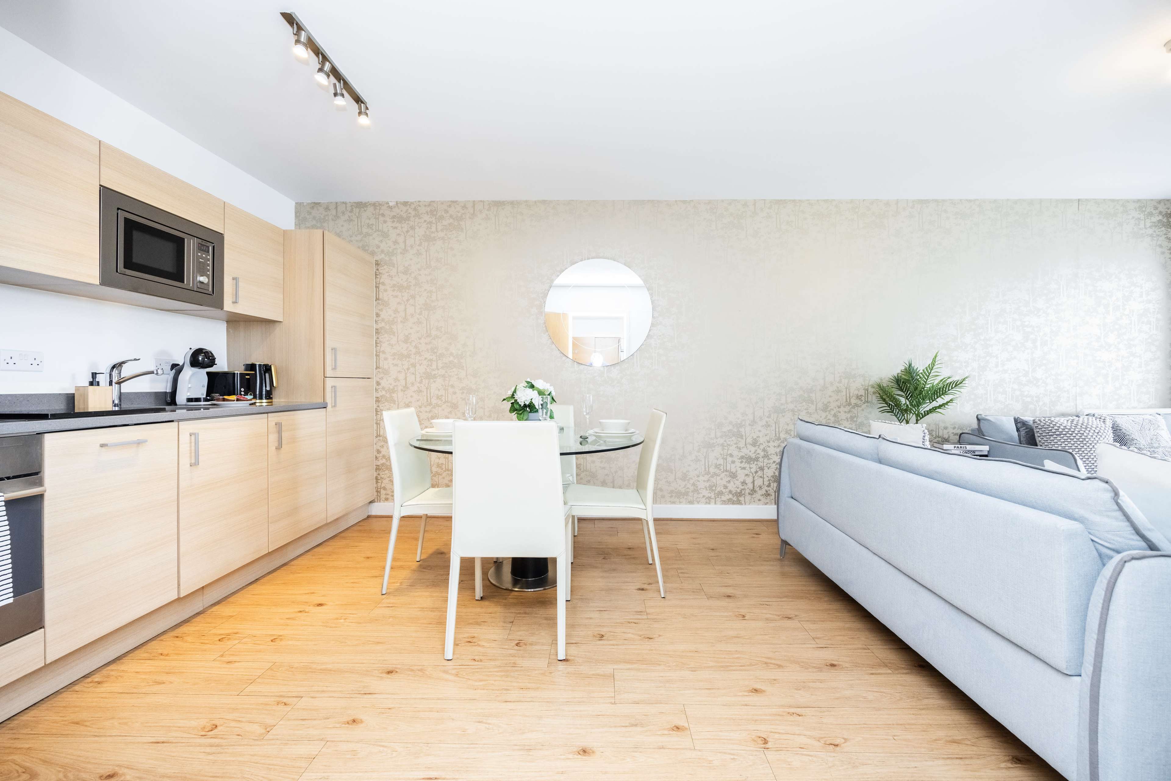 A modern kitchen and dining area with a round table, four chairs, and a living room featuring a light-colored sofa against a decorative wall.