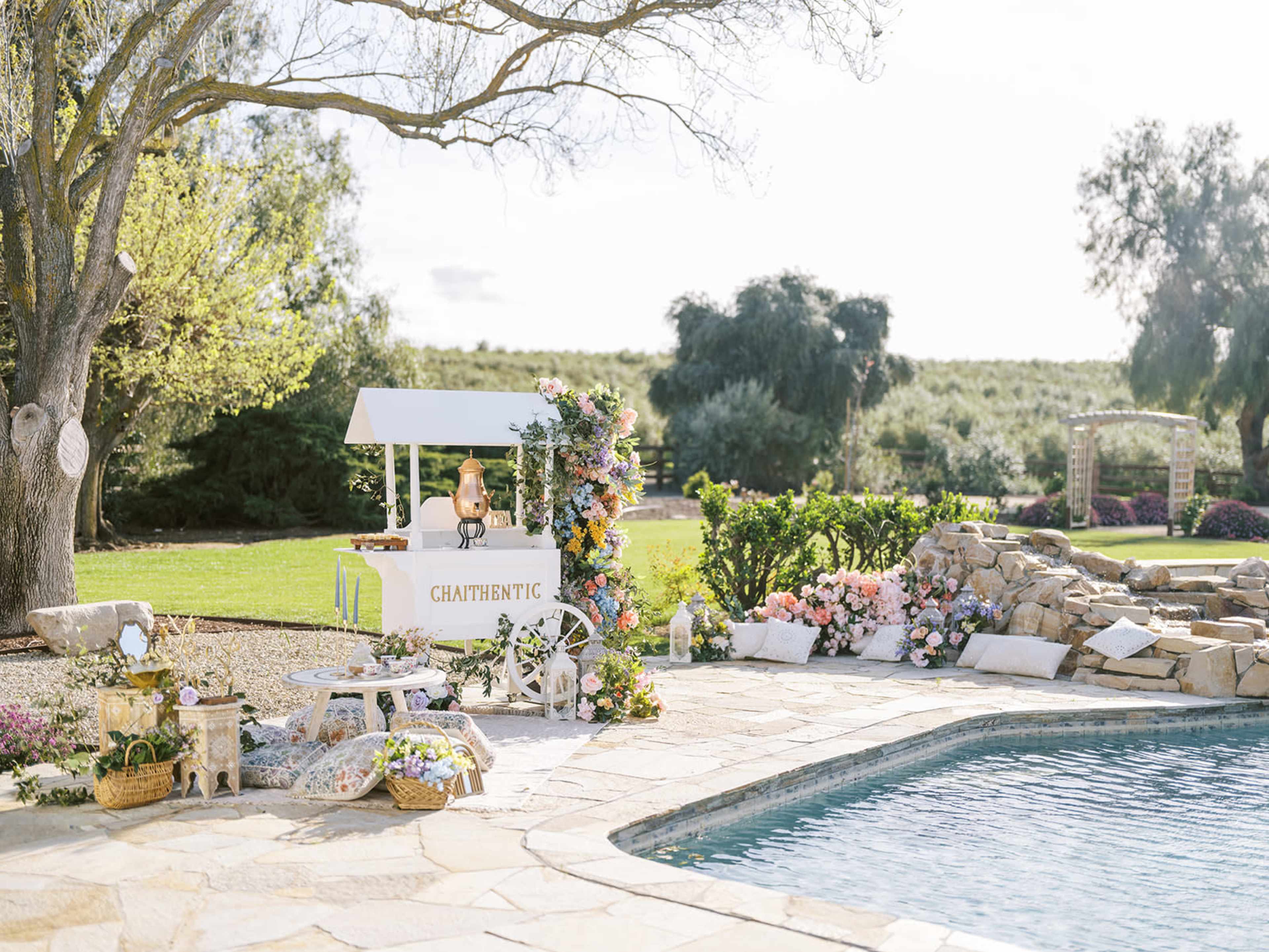 A white cart decorated with flowers stands beside a pool in a landscaped garden, with seating and various floral arrangements surrounding it.