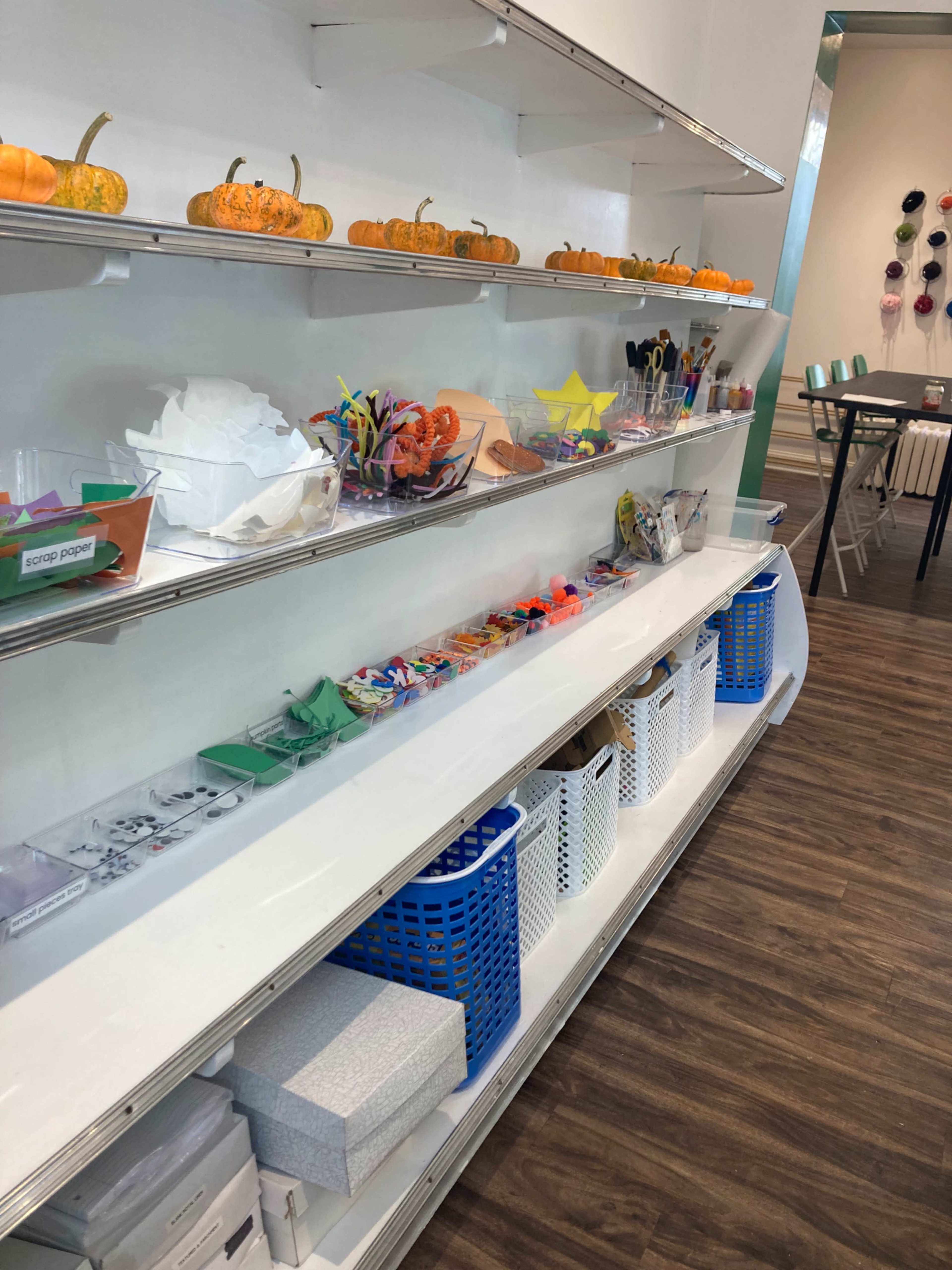 The image shows a brightly lit crafting space with organized shelves containing various crafting supplies, including small pumpkins, colorful paper, and containers filled with art materials.