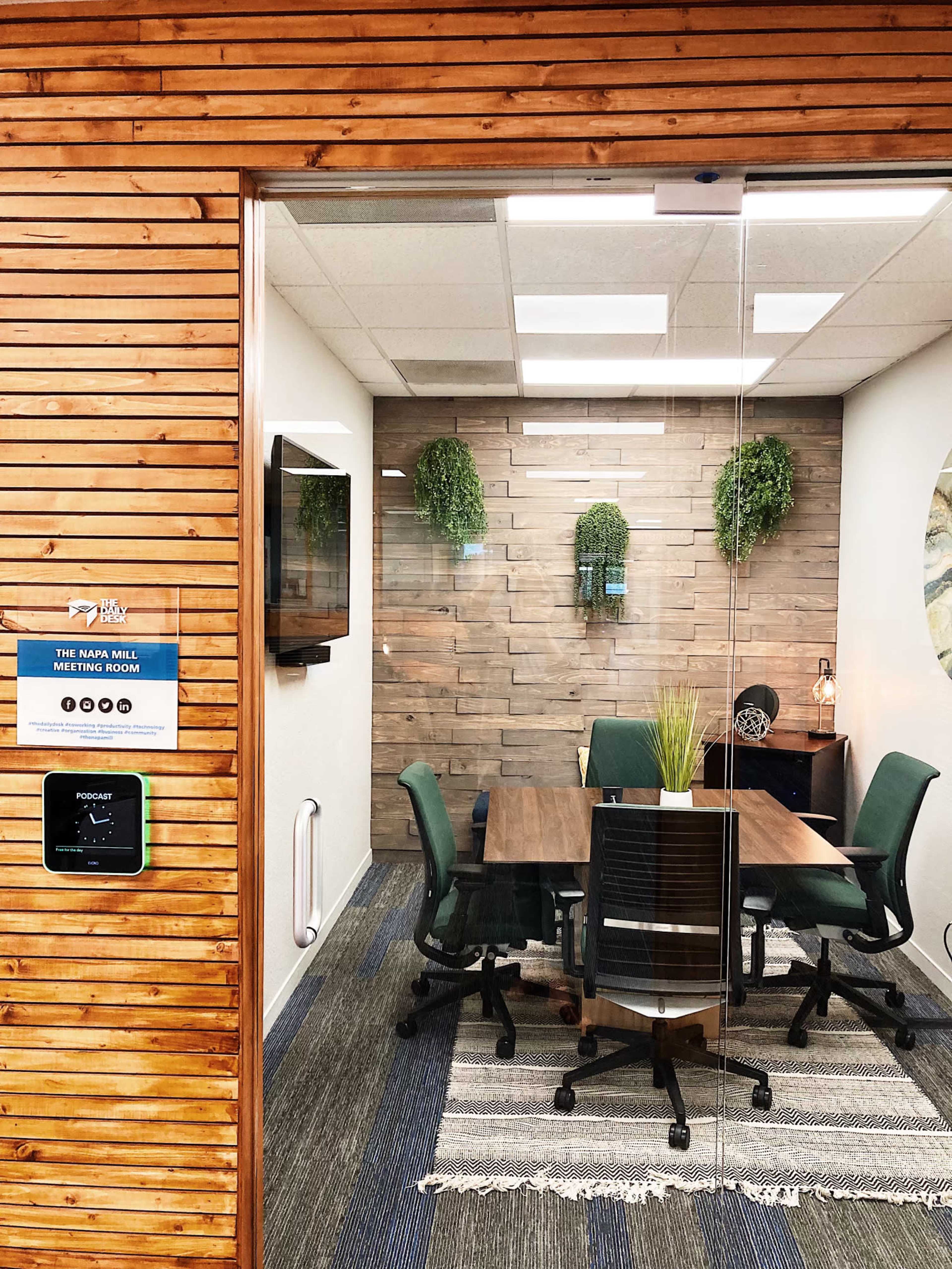 A modern meeting room with a wooden accent wall, a central table surrounded by green chairs, and decorative plants.