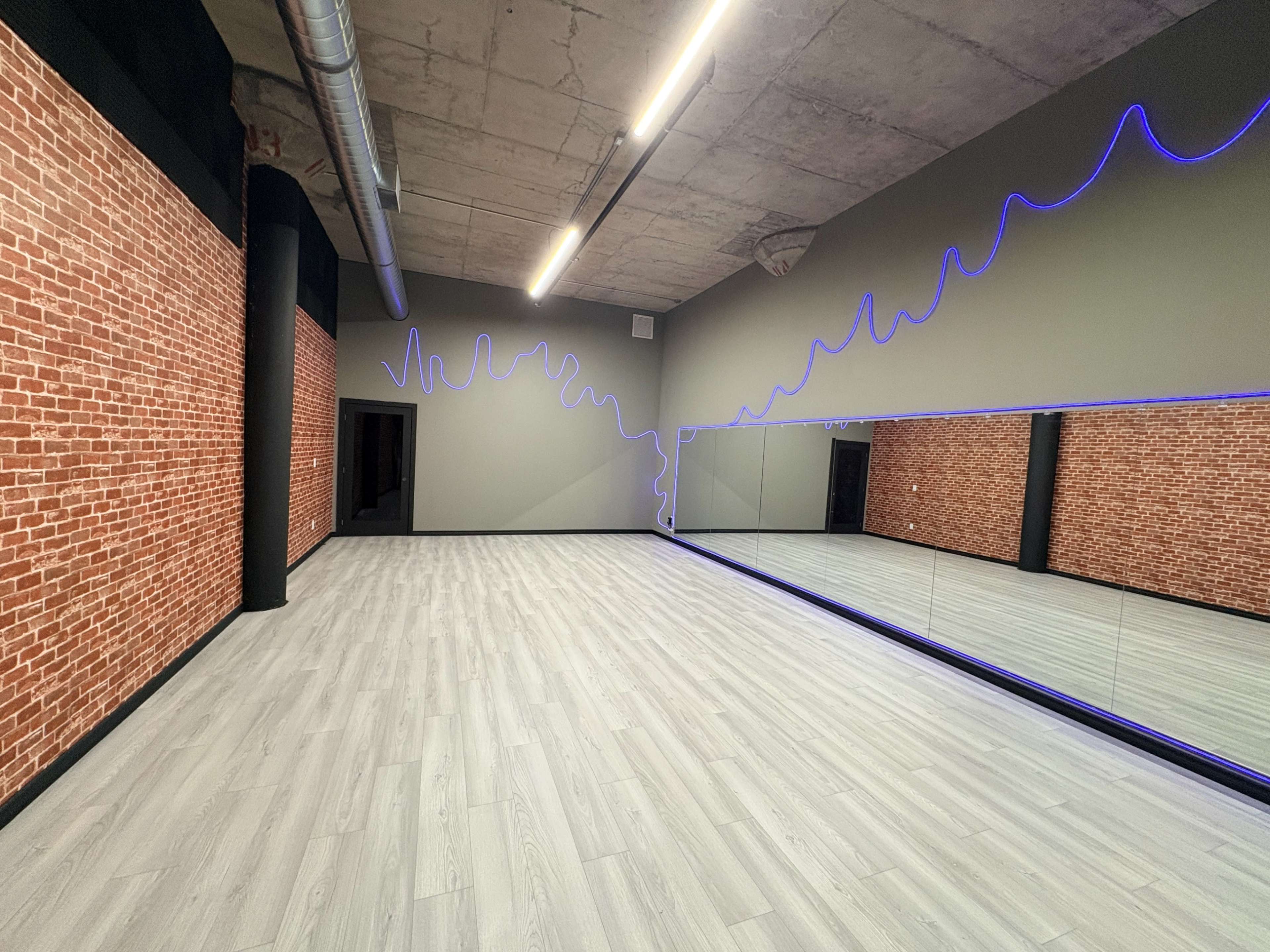 The image shows a spacious studio with exposed brick walls, a mirrored wall, and a neon light design along one wall, set in a modern, minimalistic style.