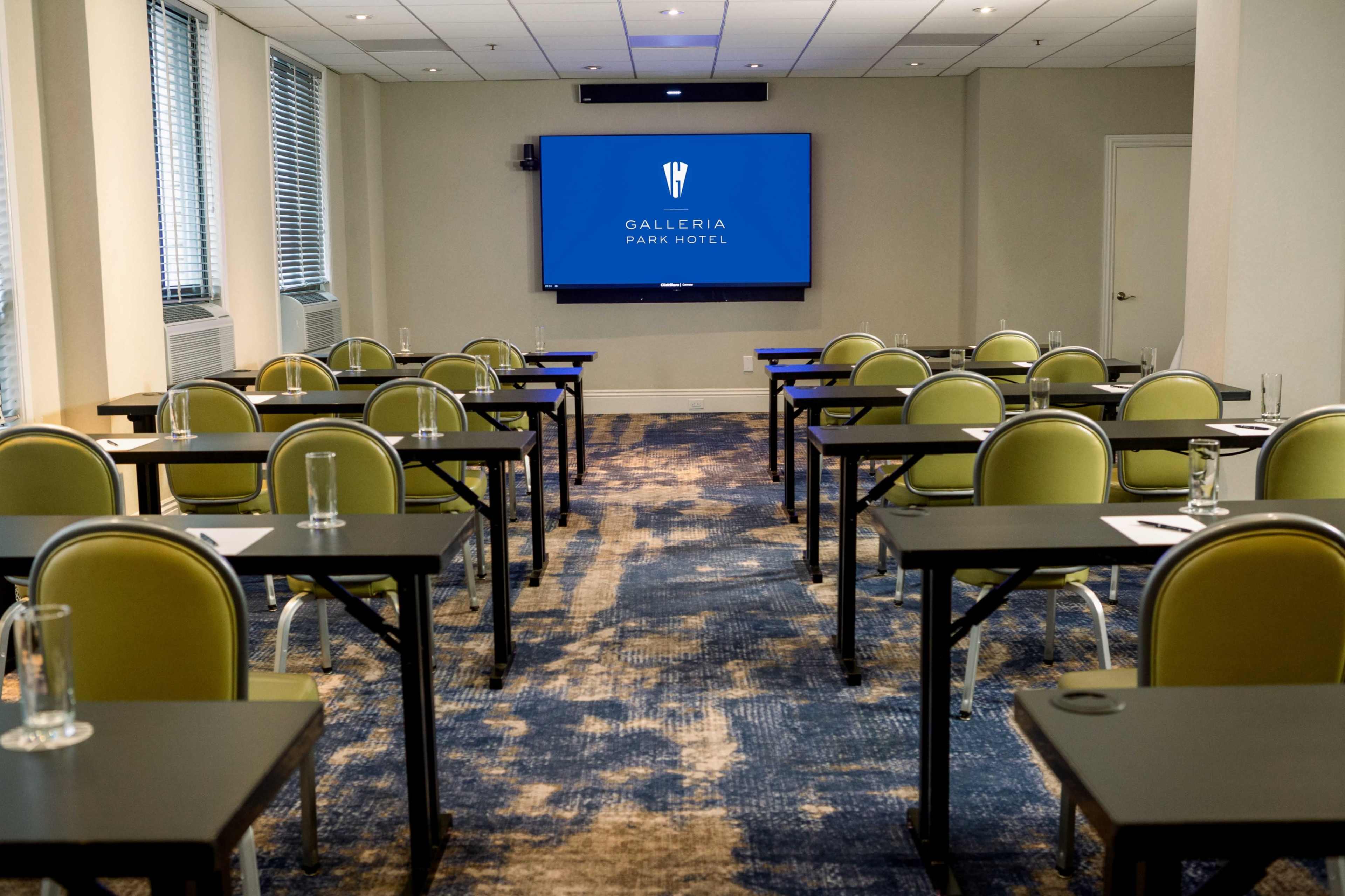 A meeting room with rows of tables and chairs facing a large screen displaying the "Galleria Park Hotel" logo.
