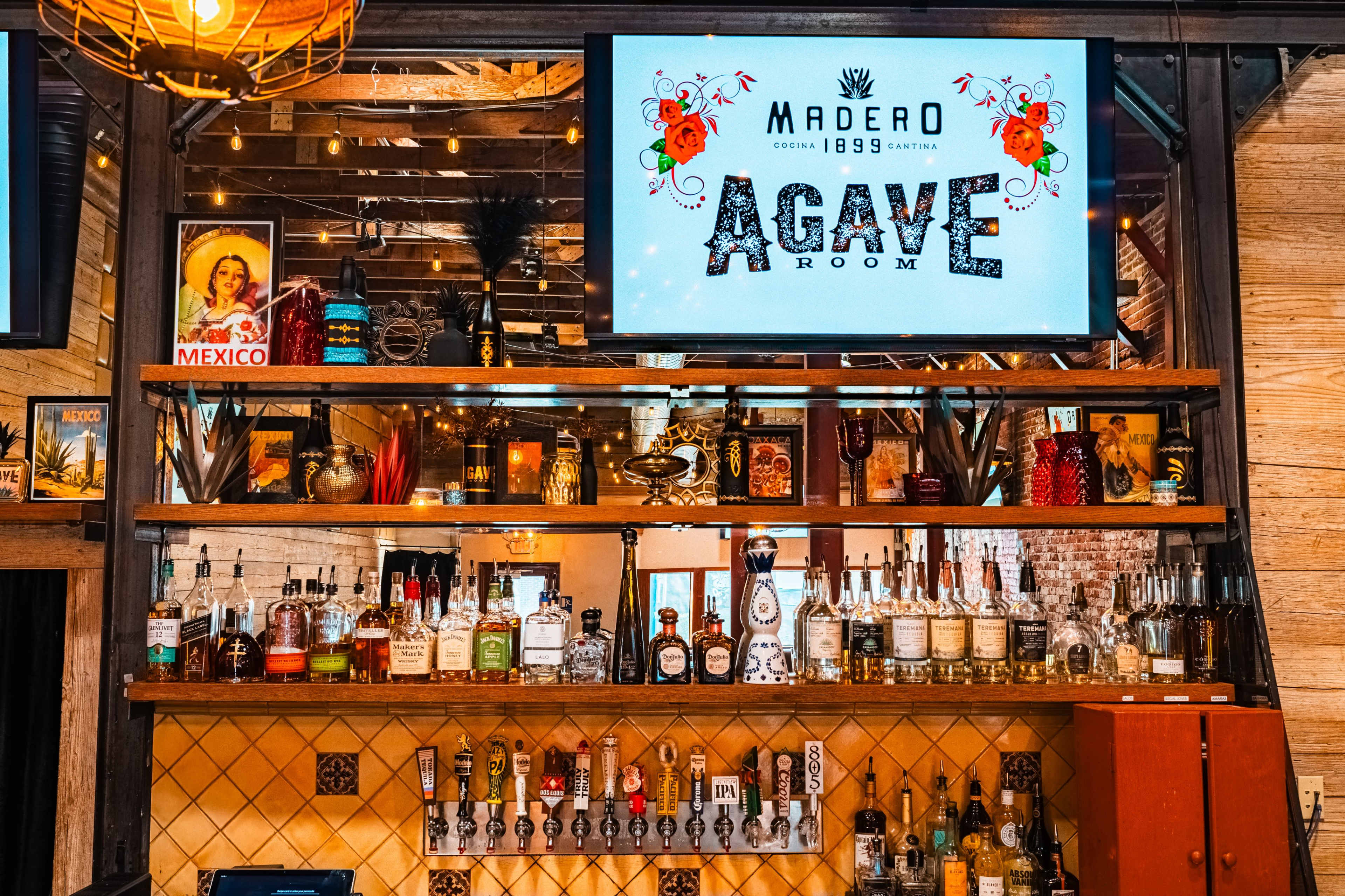 The image shows a bar area with a large sign reading "AGAVE ROOM," surrounded by shelves stocked with various bottles of alcohol and decorative items.