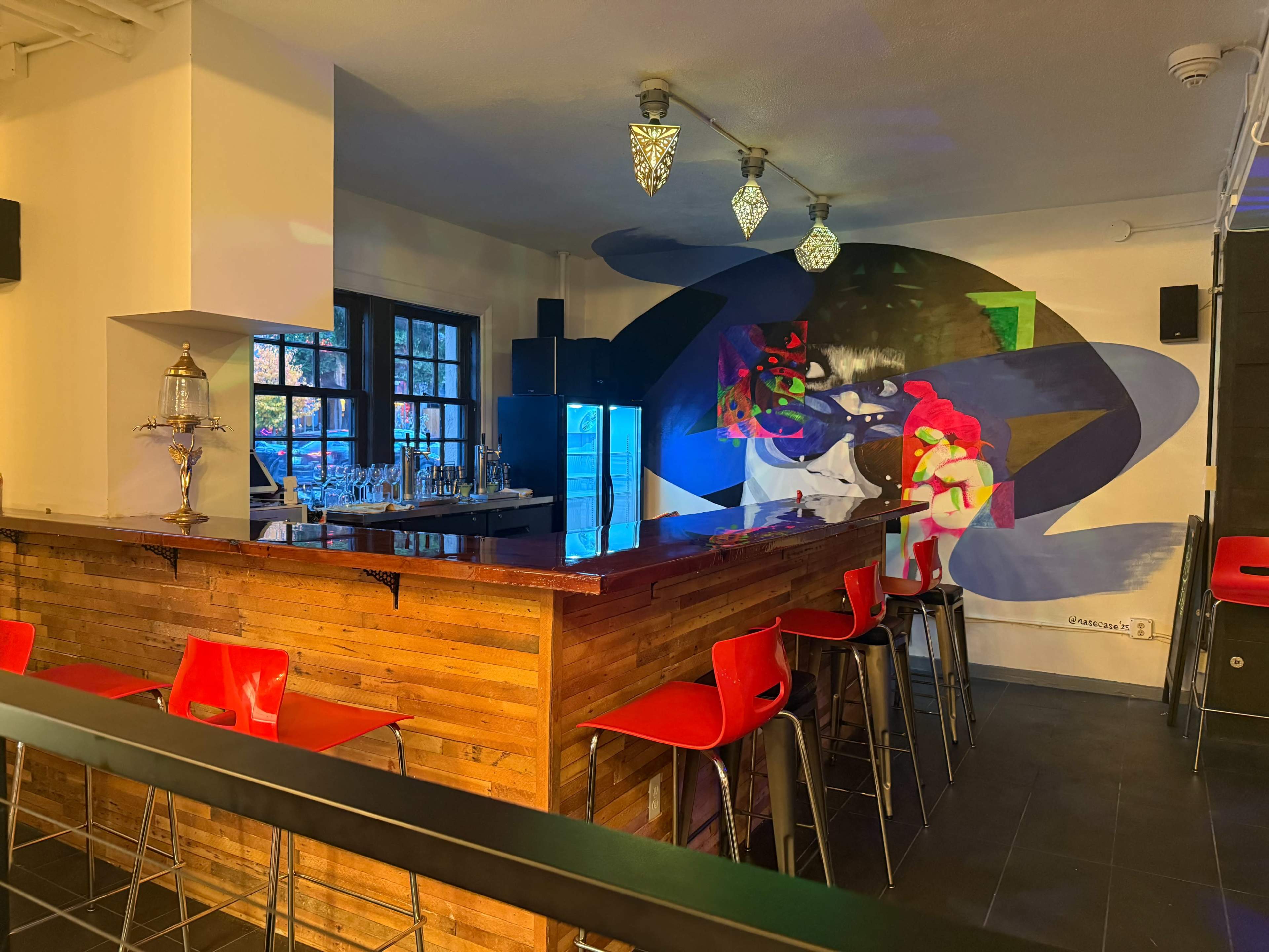 The image shows a modern bar area featuring a wooden counter, red stools, and a colorful abstract mural on the wall.