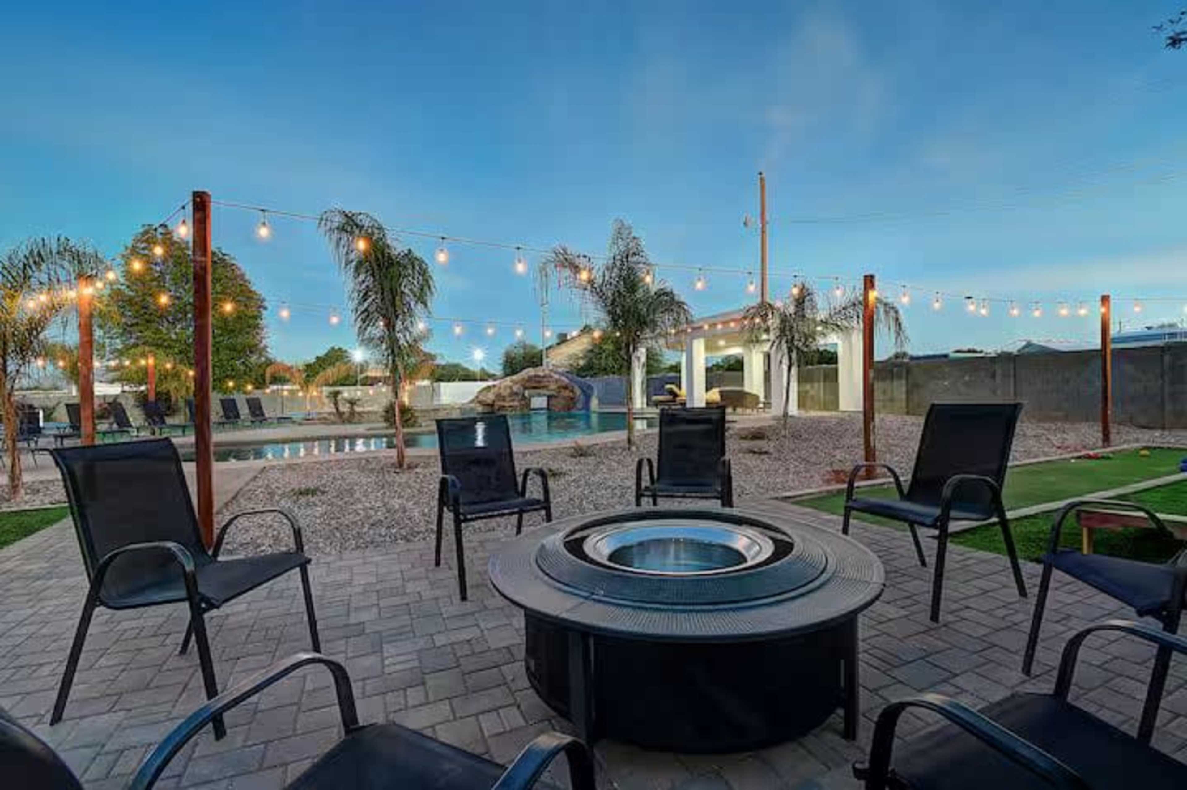 The image shows a backyard patio area with a fire pit surrounded by chairs, leading to a pool and landscaped yard illuminated by string lights.