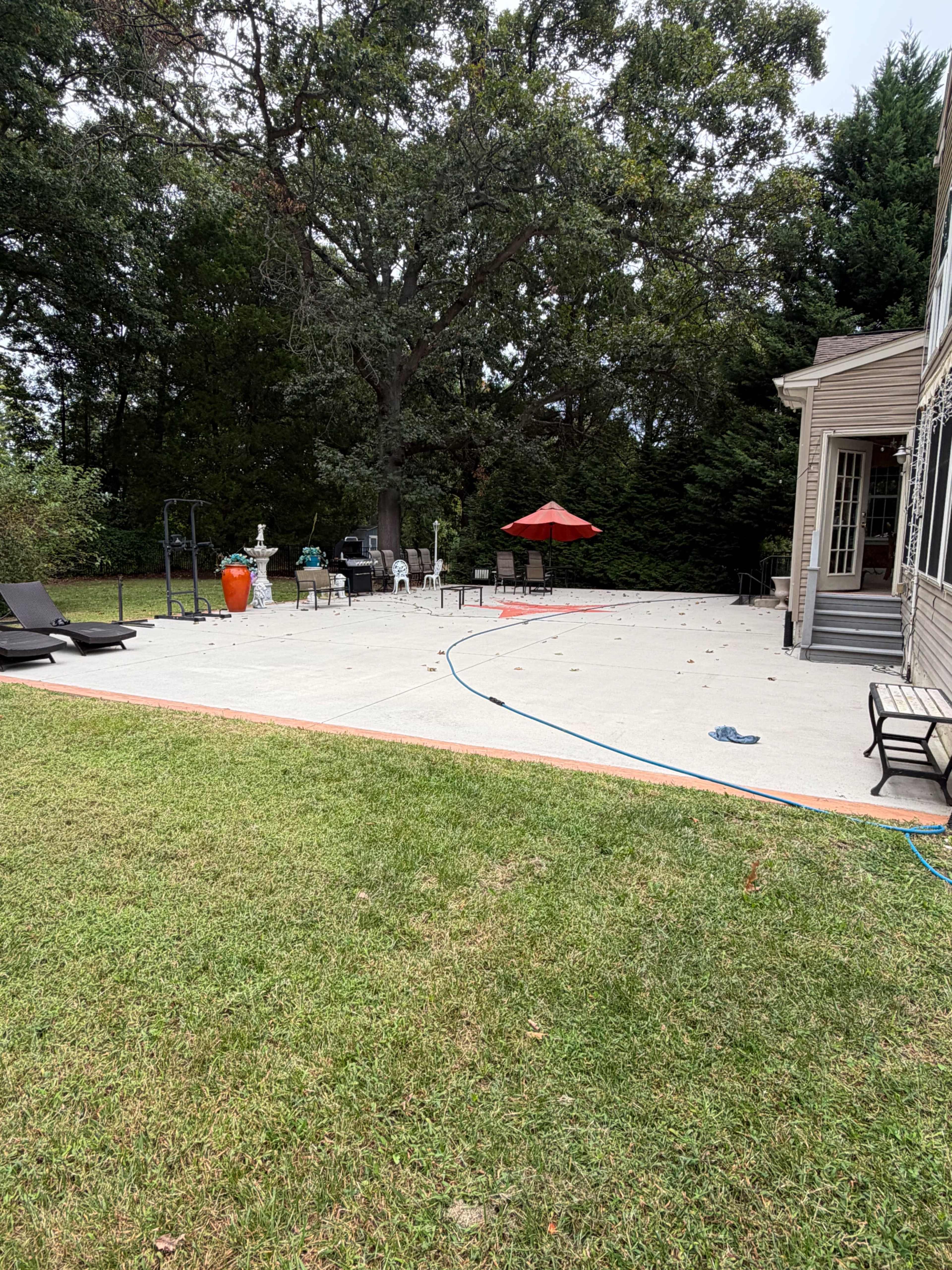 The image shows a backyard patio area with lounge chairs, a red umbrella, and various outdoor furniture against a backdrop of trees.