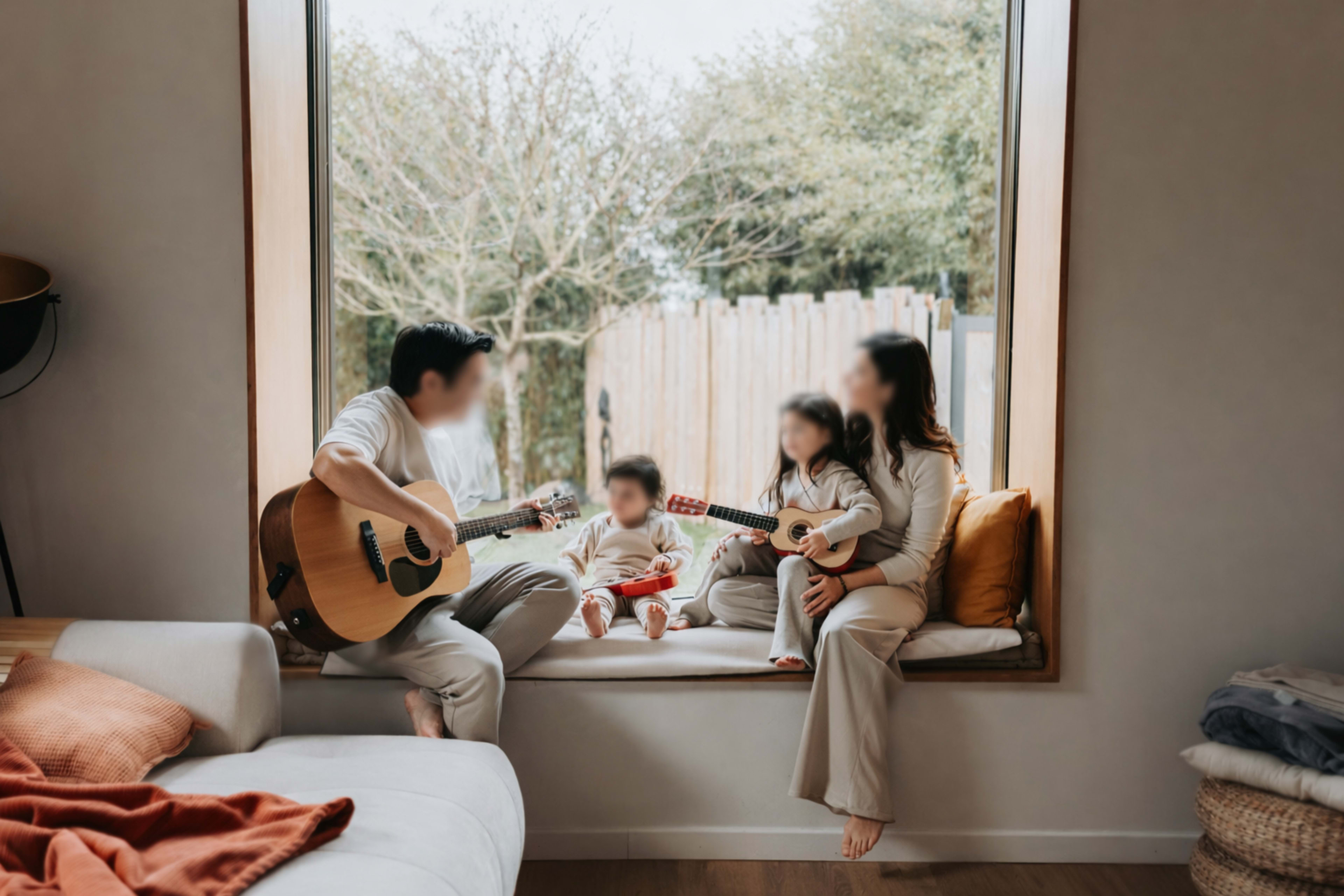 A family of four sits in a window nook, playing guitars and enjoying time together in a cozy living room.