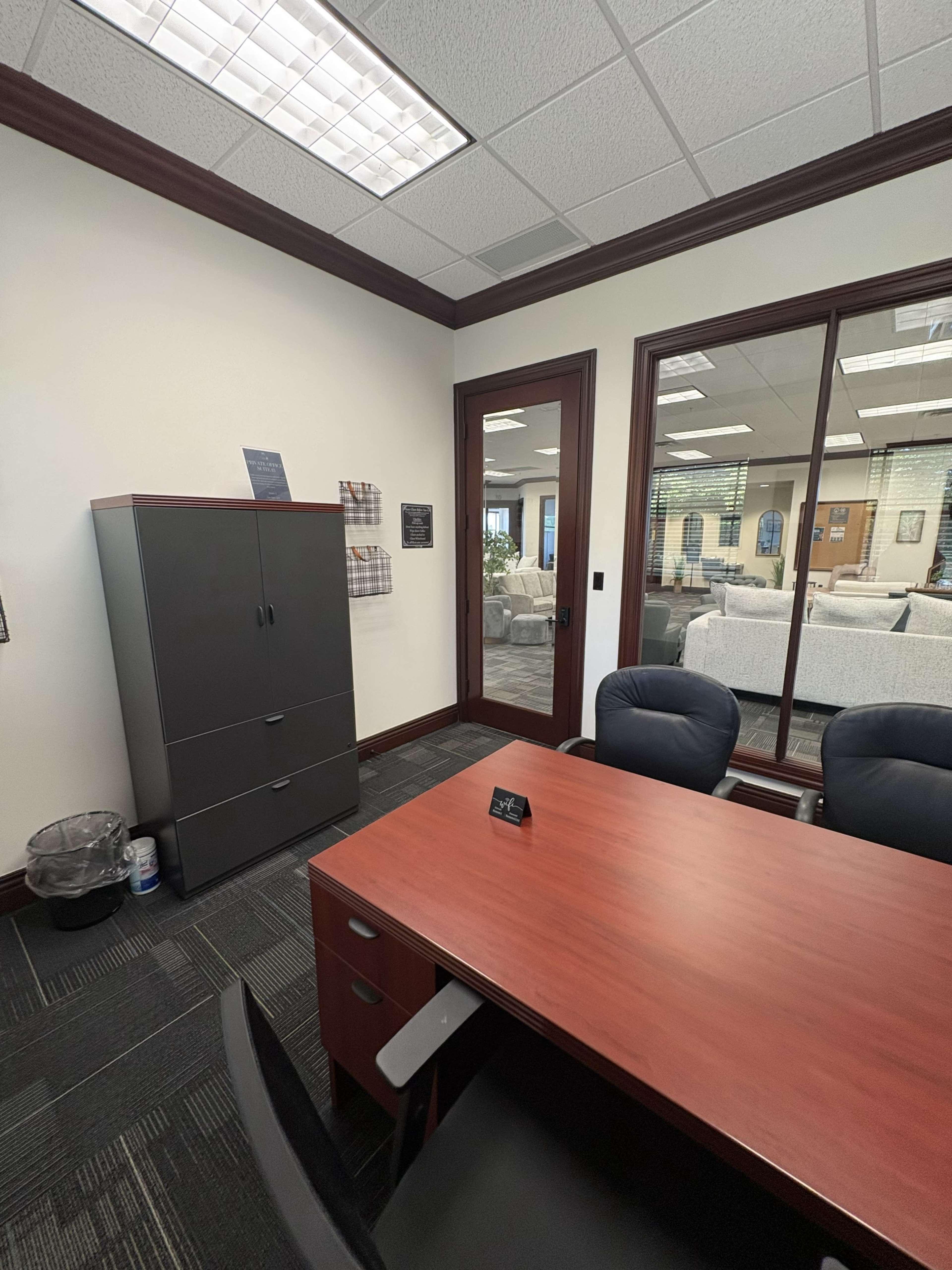 The image shows a modern office space featuring a wooden desk and black chairs, with a grey filing cabinet and a large window overlooking a seating area.
