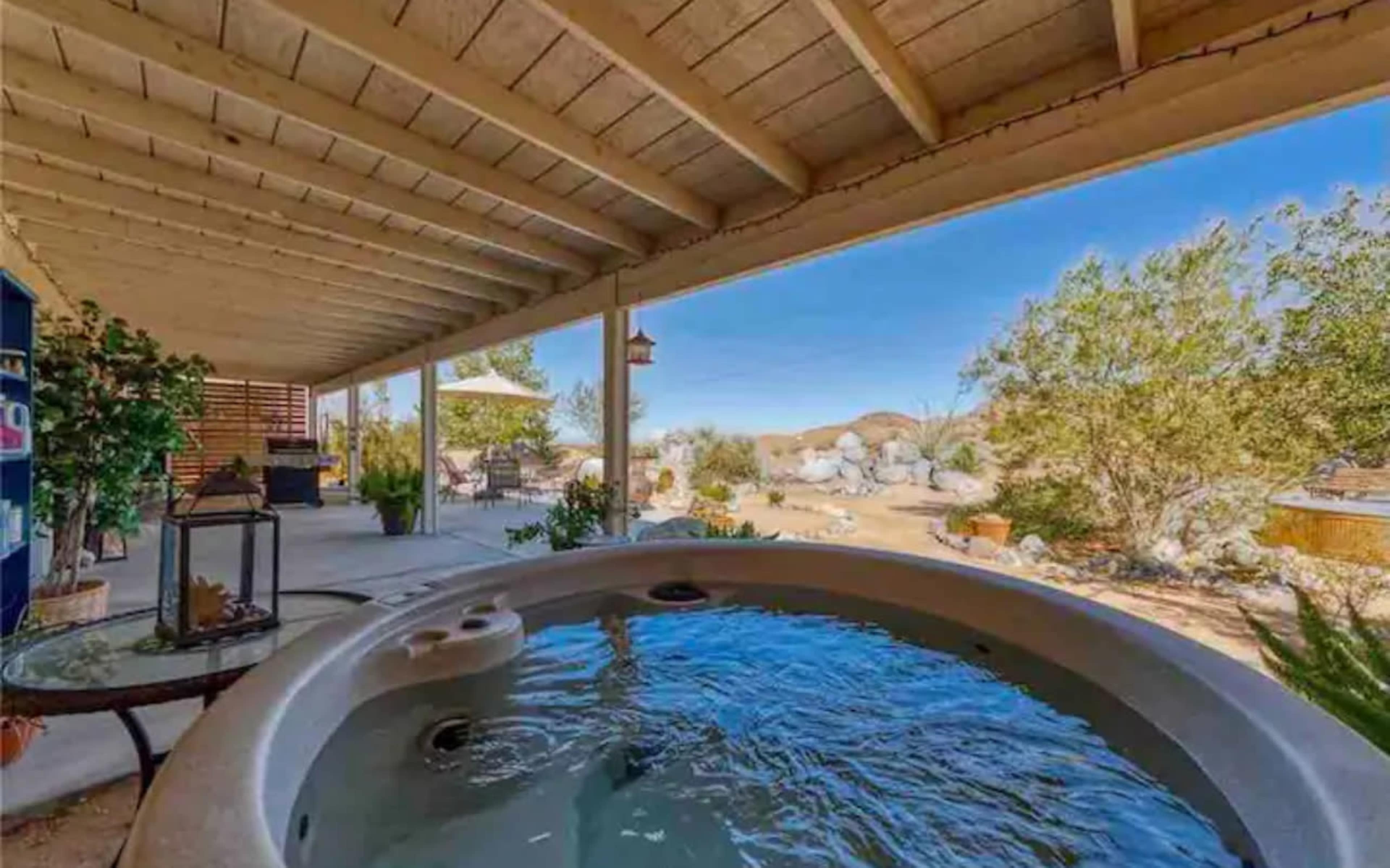 A hot tub is positioned under a covered patio, overlooking a desert landscape with rocky formations and sparse vegetation.